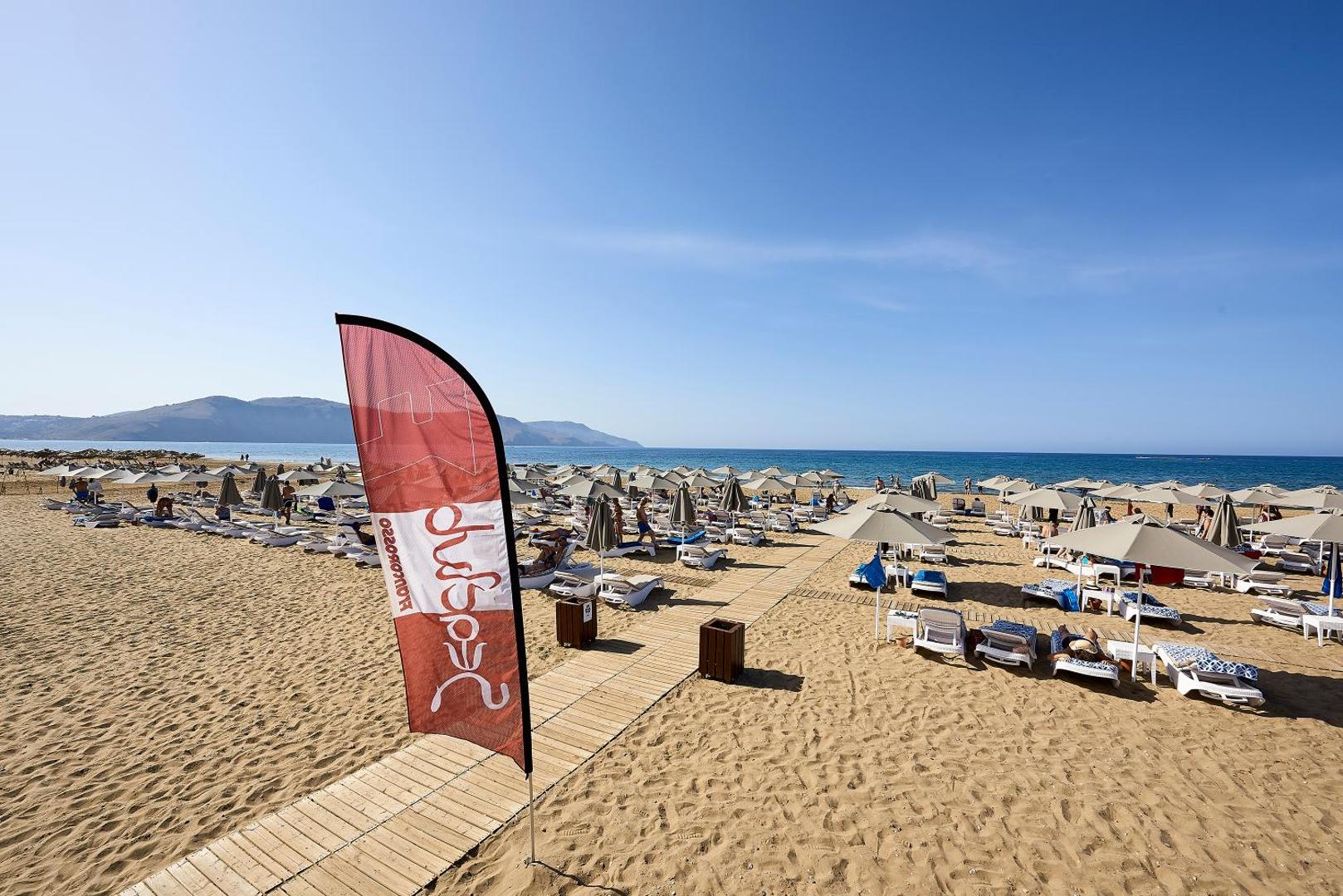 Mythos Palace Resort & Spa plašā smilšu pludmale Georgiupoli, Krētā ar saulessargiem, zviļņiem un koka laipām pie dzidras jūras