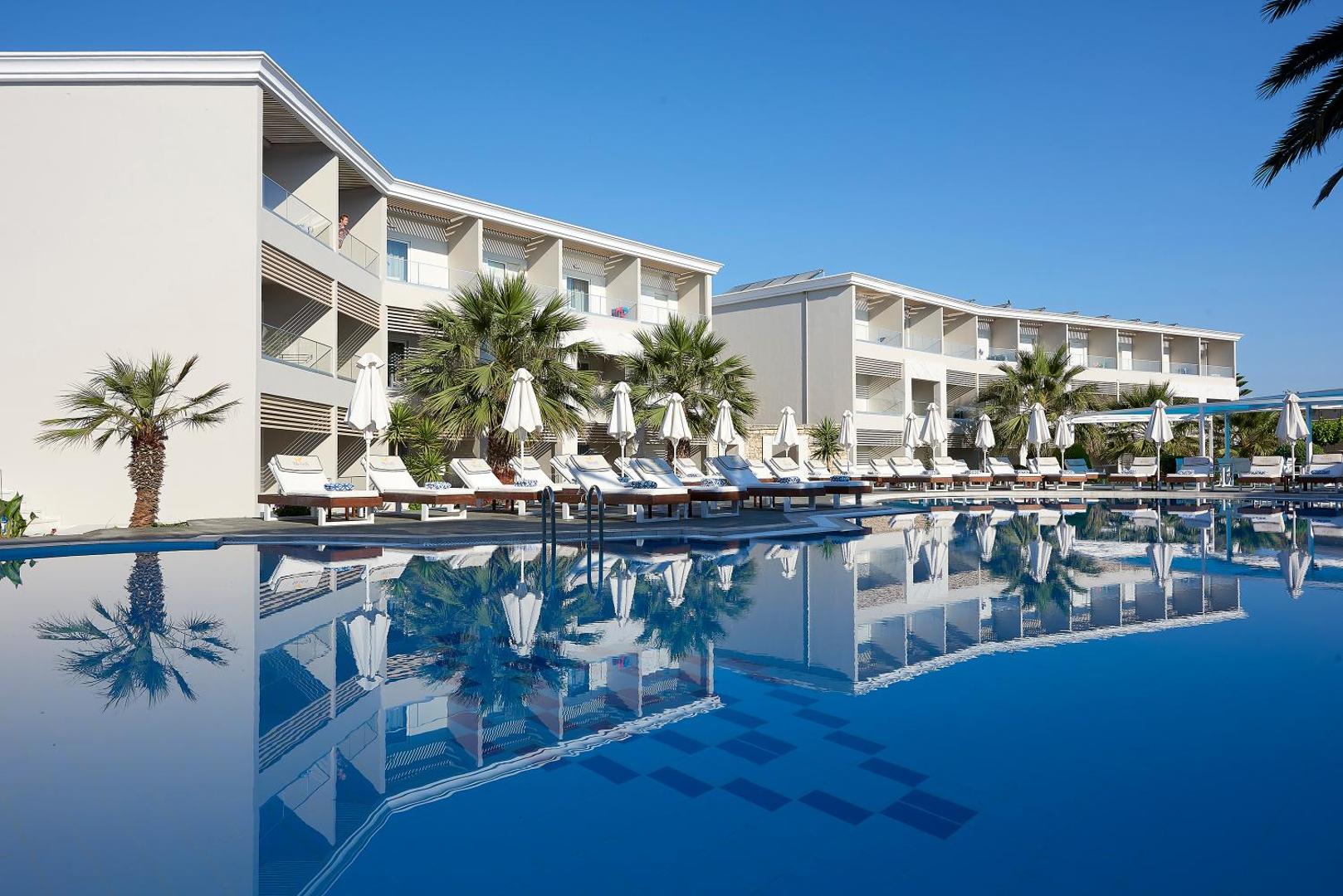 Mythos Palace Resort & Spa modernie numuru korpusi pie liela āra baseina ar saulessargiem un palmām dienas laikā