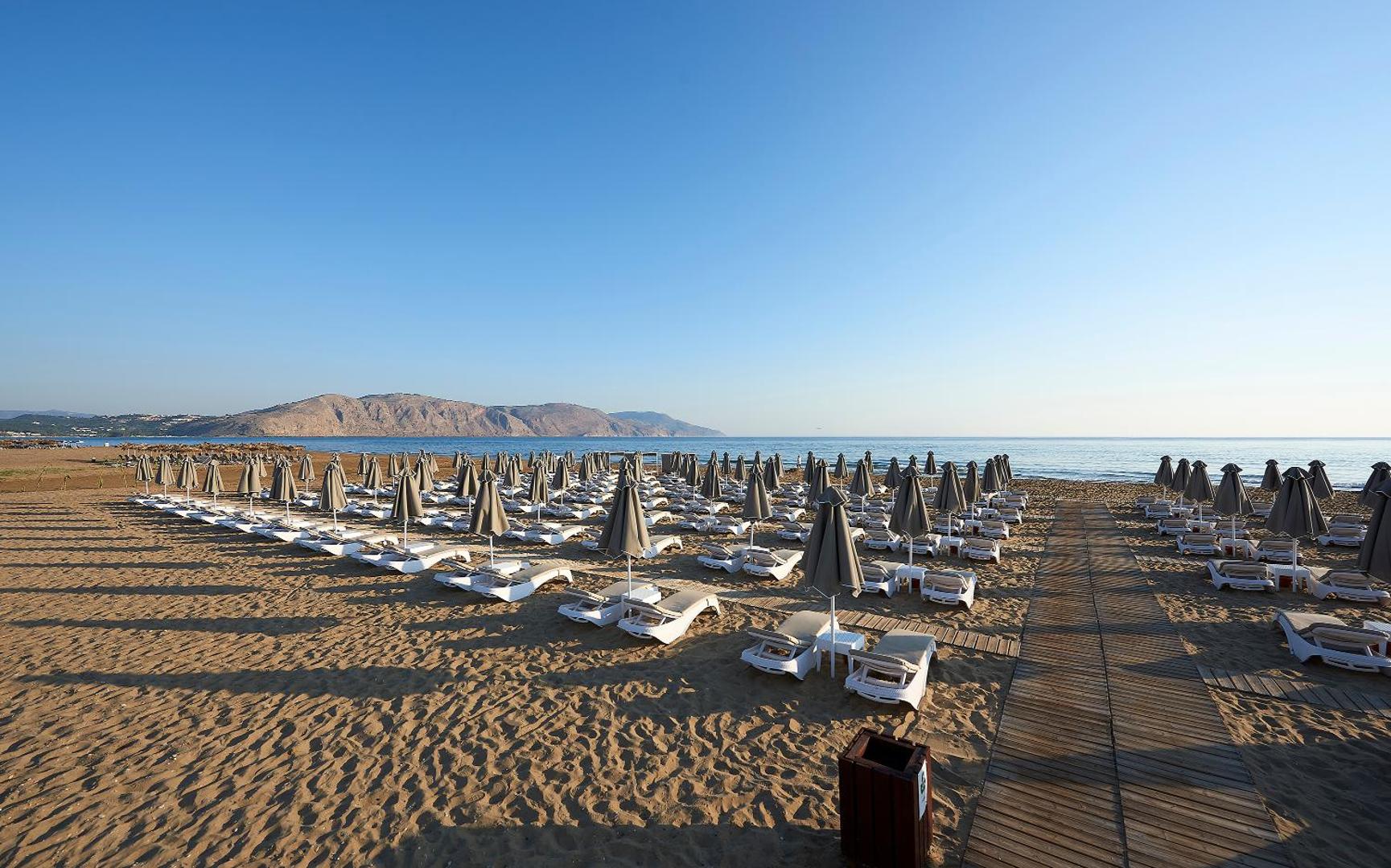 Mythos Palace Resort & Spa privātā pludmales zona rīta gaismā ar sakārtotām sauļošanās vietām un skatu uz kalniem