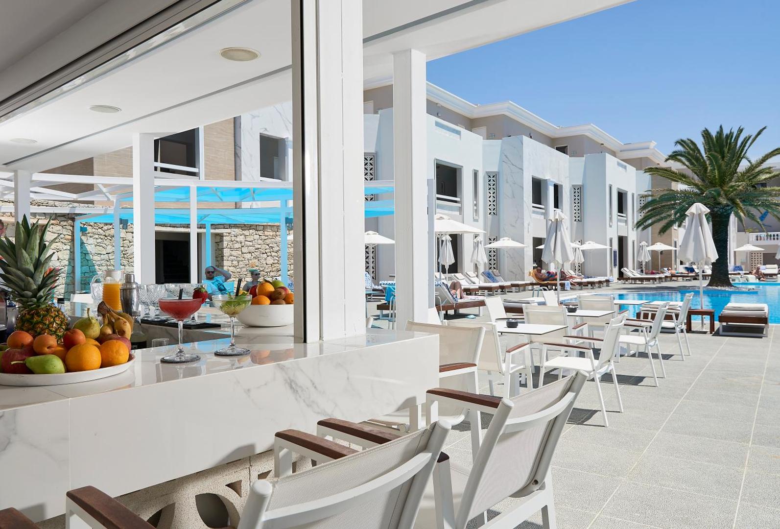Mythos Palace Resort & Spa āra bārs ar augļiem un kokteiļiem priekšplānā, skats uz baseina zonu un viesnīcas ēkām