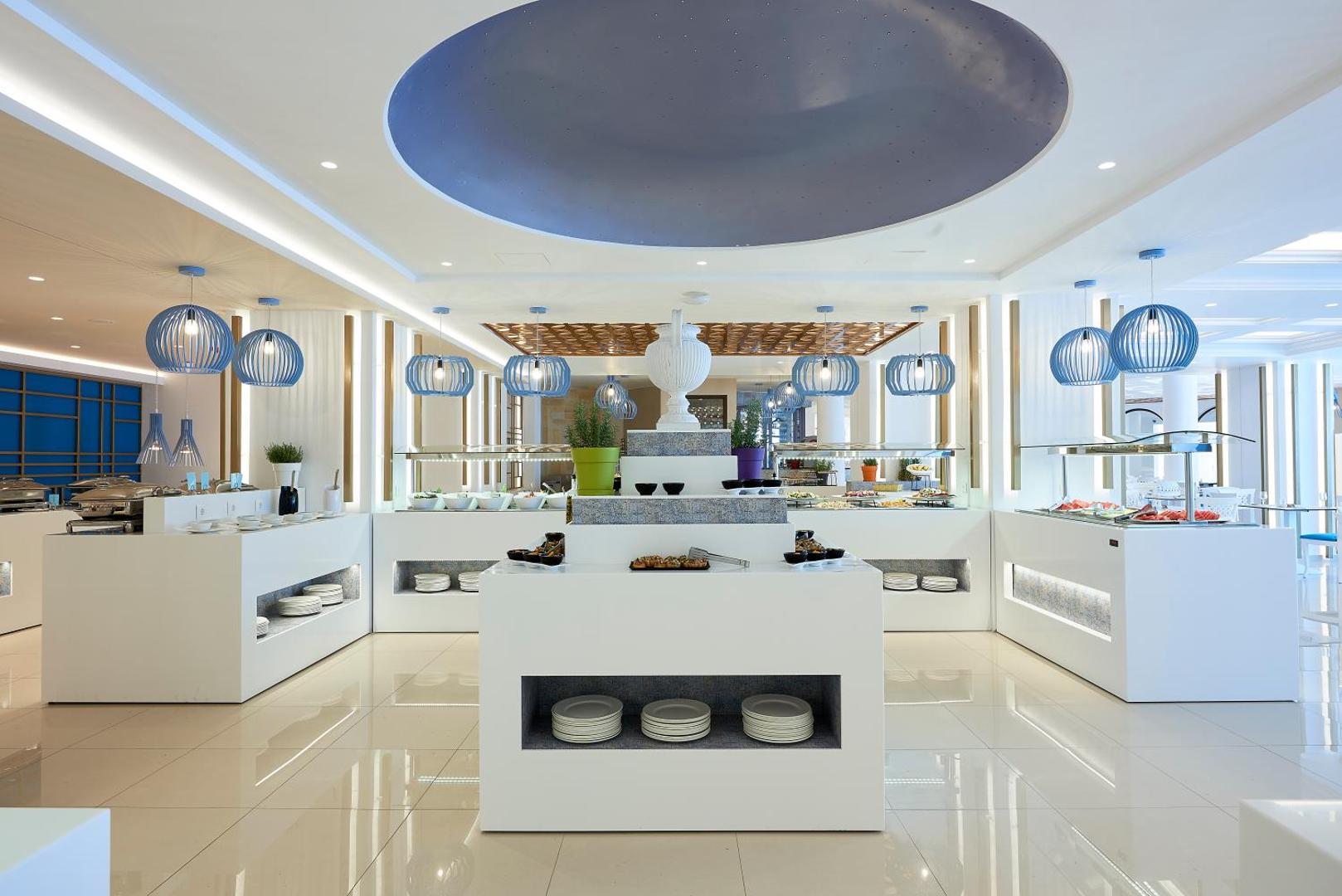 Mythos Palace Resort & Spa restorāna bufetes zona ar modernu dizainu, baltām mēbelēm un plašu ēdienu izvēli