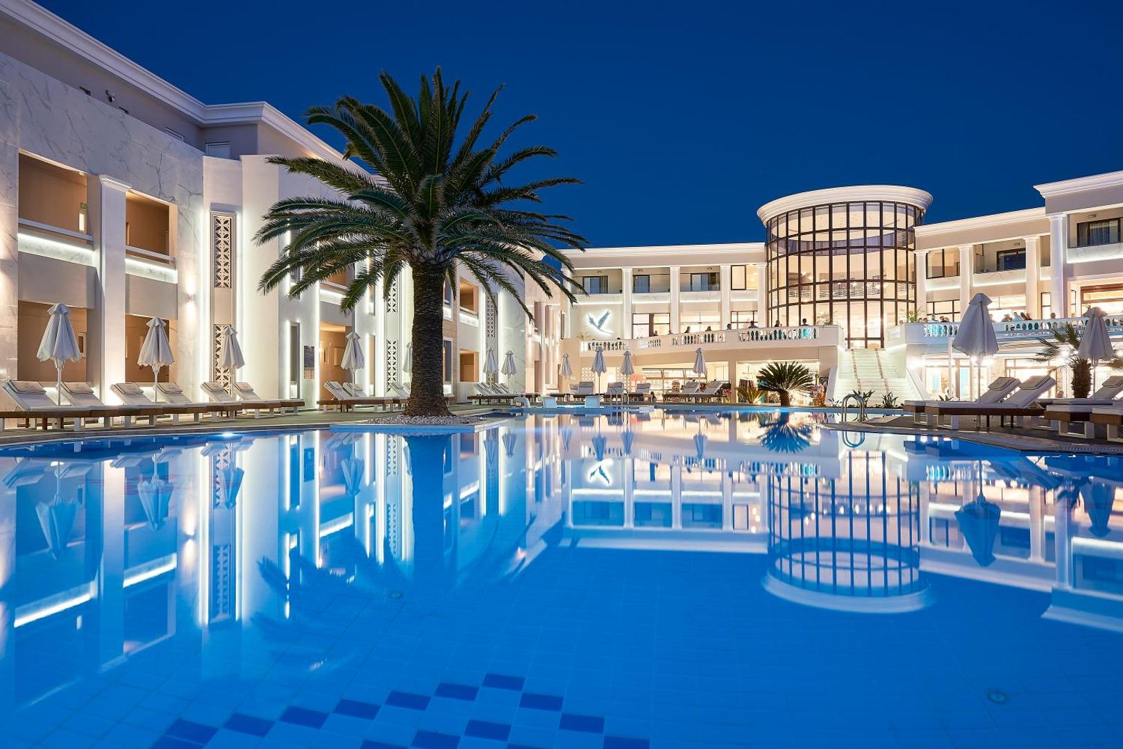 Mythos Palace Resort & Spa galvenais baseins vakarā — izgaismota viesnīcas fasāde, palmas un atpūtas krēsli pie ūdens Krētā