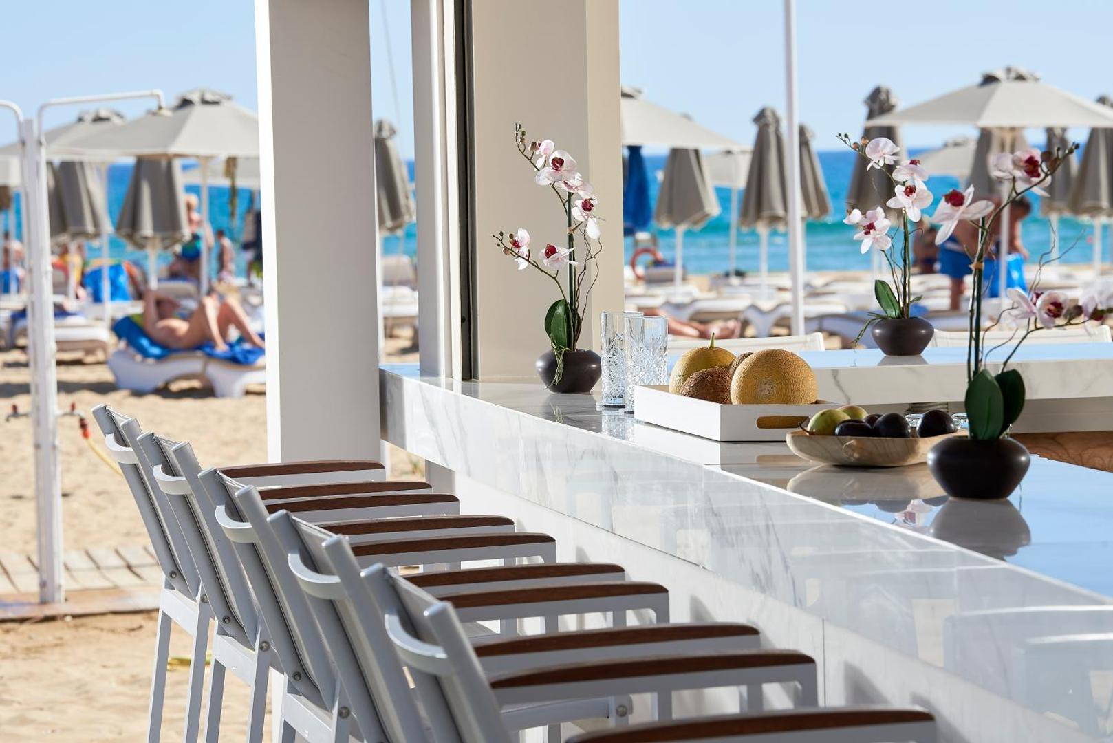 Mythos Palace Resort & Spa pludmales bārs ar skatu uz jūru, baltiem bāra krēsliem un svaigiem augļiem uz letes