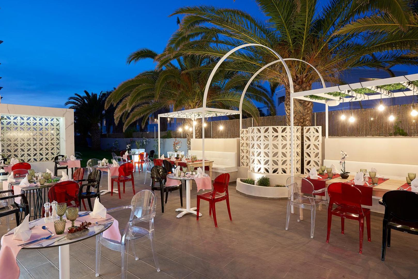 Mythos Palace Resort & Spa restorāna āra zona ar palmām un dekoratīvām arkām vakara apgaismojumā Krētas piekrastē