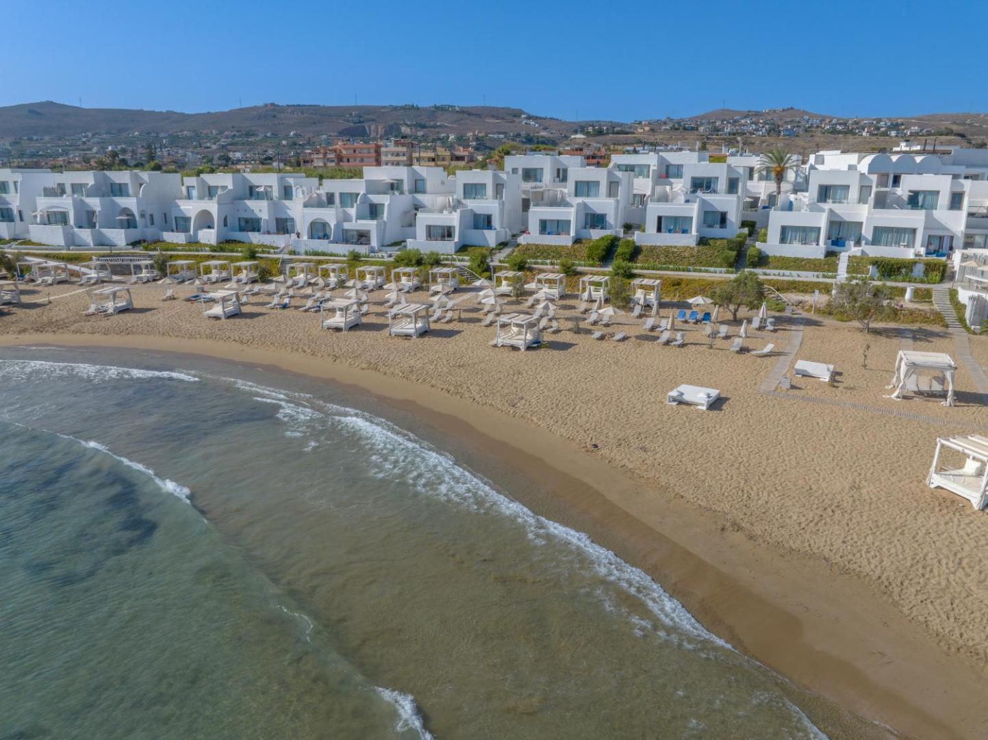 Skats no jūras puses uz Knossos Beach Bungalows Resort balto arhitektūru un labiekārtoto pludmali ar saulessargiem un atpūtas zonām