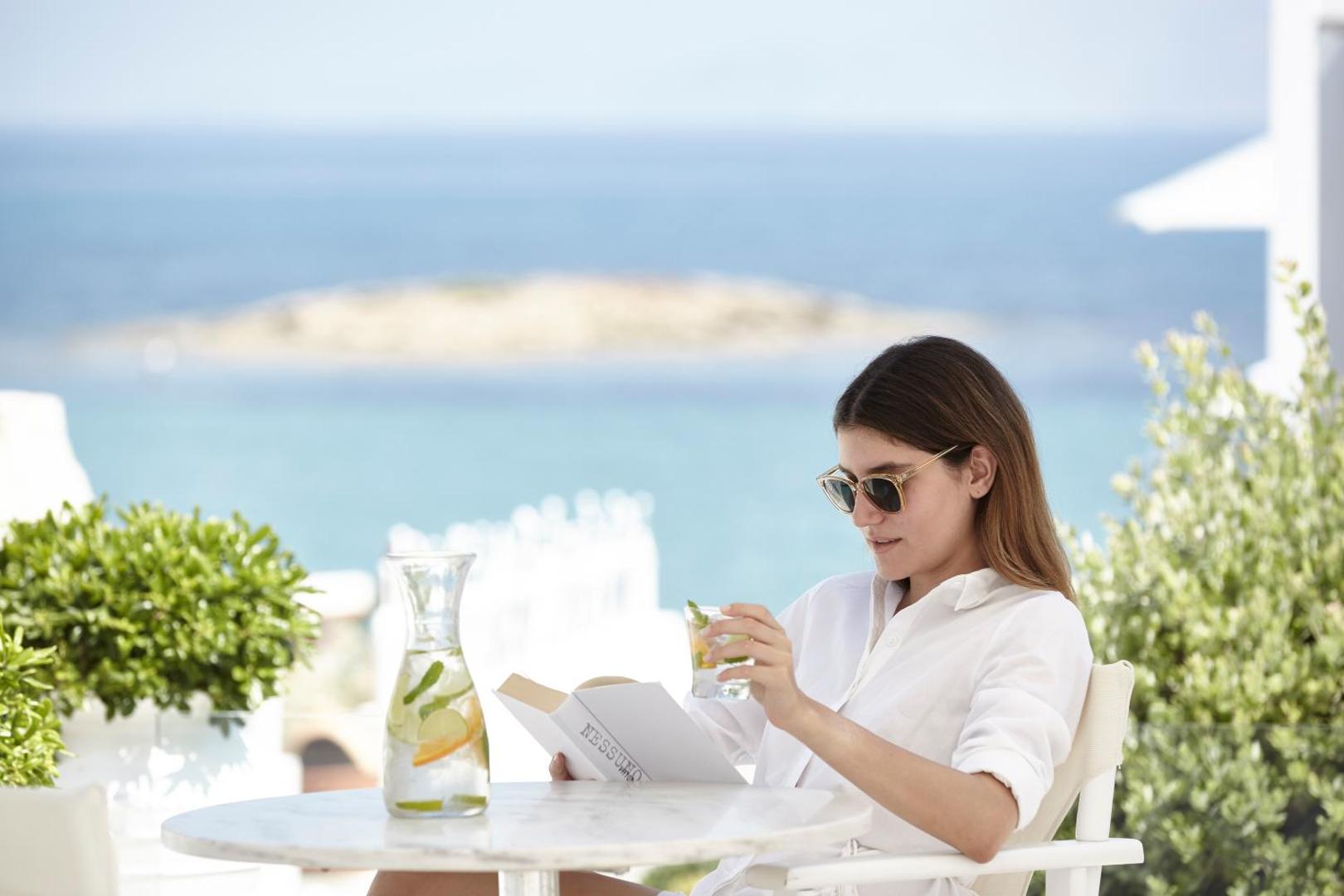 Viesis bauda atspirdzinošu dzērienu un lasa grāmatu uz Knossos Beach Bungalows Resort terases ar skatu uz Vidusjūru