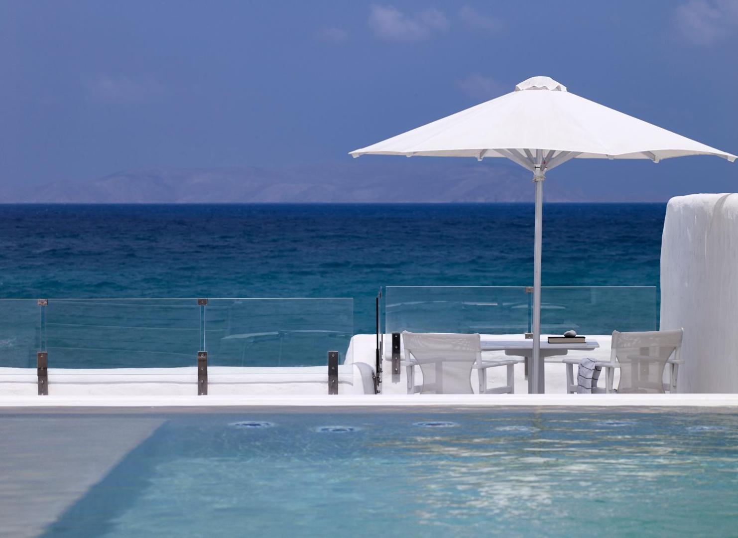 Knossos Beach Bungalows Resort terase ar saulessargu un galdiņu pie baseina malas, fonā tumši zilā Egejas jūra