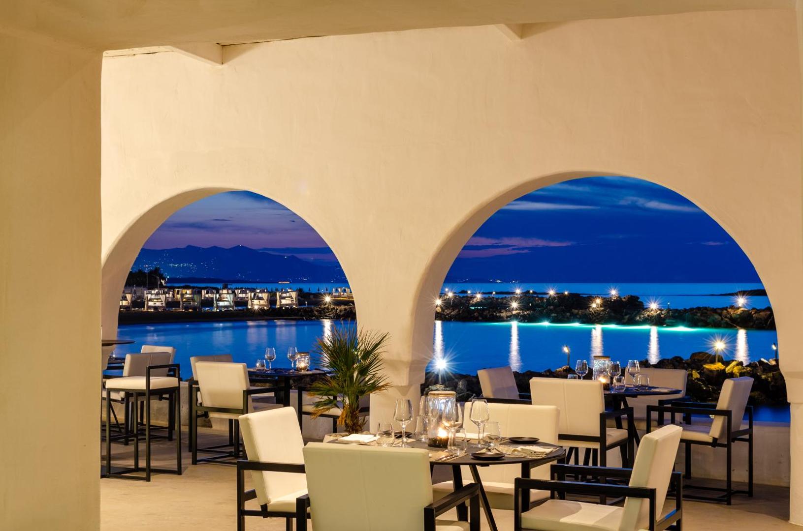 Knossos Beach Bungalows Resort restorāna terase vakarā ar izgaismotām arkām un skatu uz izgaismoto piekrasti un kalniem
