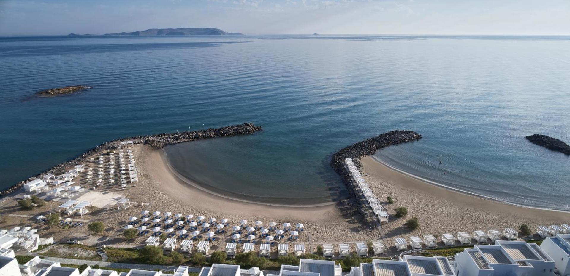Knossos Beach Bungalows Resort panorāmas skats uz privāto smilšu pludmali un līci Krētā ar saulessargiem un atpūtas krēsliem pie jūras