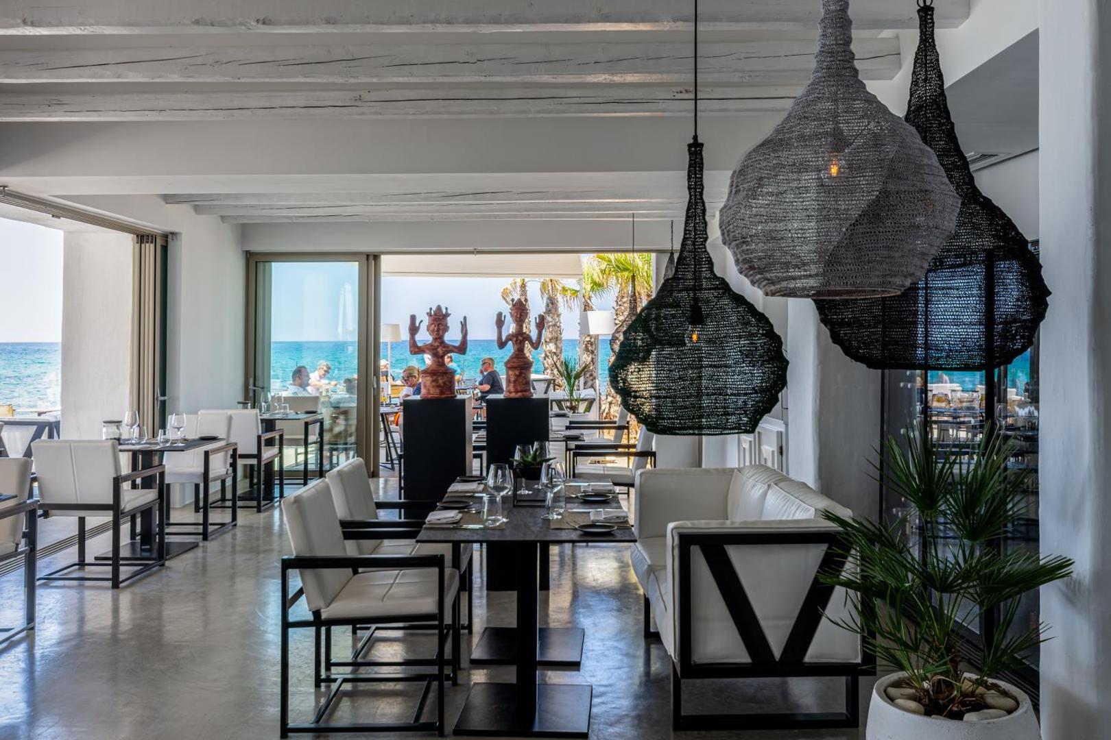 Knossos Beach Bungalows Resort moderns restorāna interjers ar dizaina gaismekļiem un skatu uz zilo jūru caur logiem