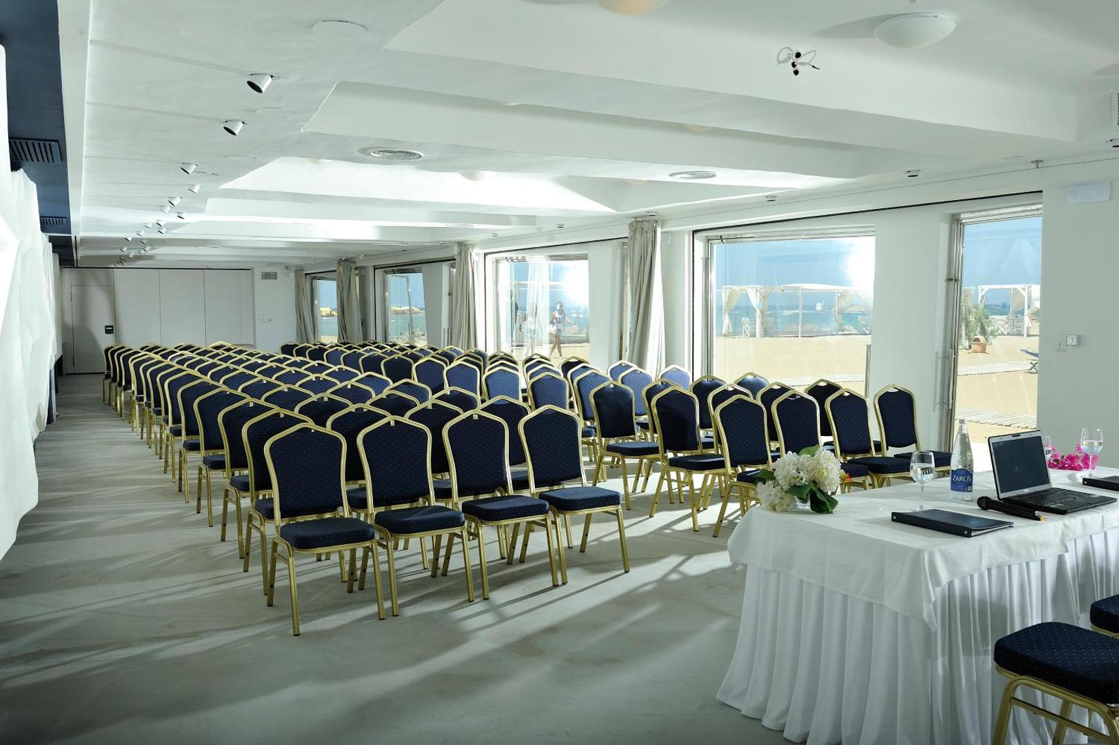 Knossos Beach Bungalows Resort konferenču zāle ar zilām krēsliem un skatu uz pludmali caur lieliem logiem