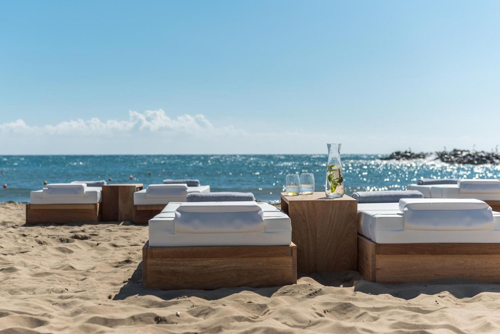 Ekskluzīvas koka atpūtas gultas ar baltiem matračiem Knossos Beach Bungalows Resort pludmalē ar skatu uz zilo jūru un atsvaidzinošiem dzērieniem