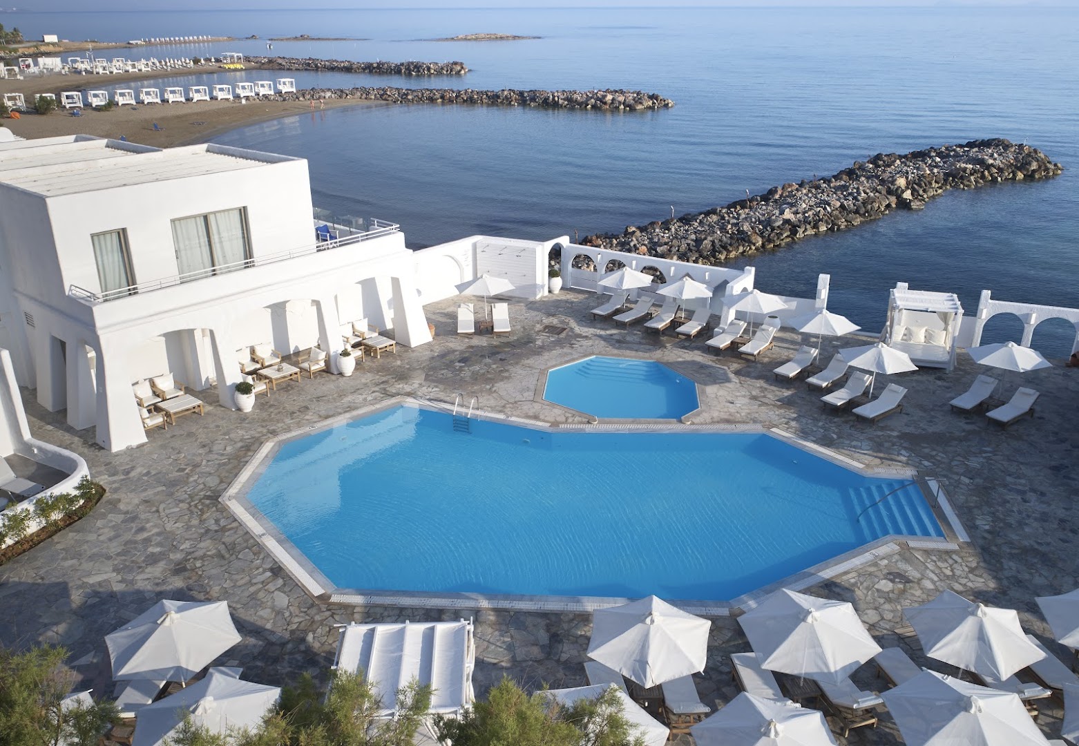 Knossos Beach Bungalows Resort panorāmas skats uz baseiniem un privāto pludmali Krētā, baltas ēkas un sauļošanās krēsli piekrastē
