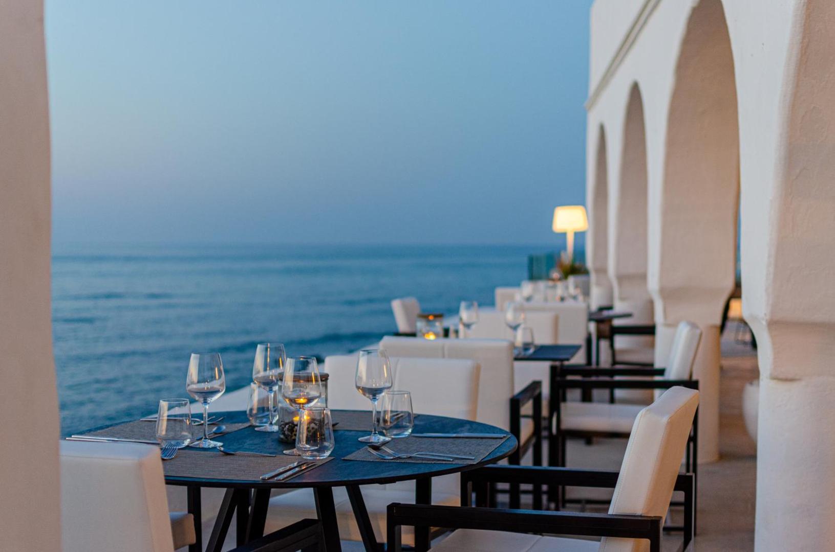 Knossos Beach Bungalows Resort restorāna āra terase ar klātiem galdiem tieši pie jūras krasta līnijas saulrietā