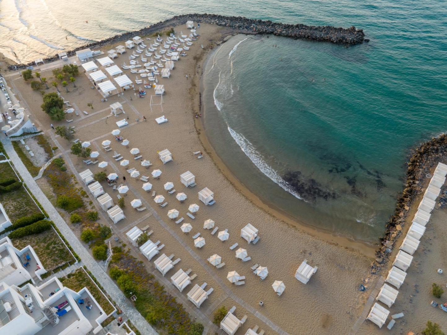Knossos Beach Bungalows Resort pludmales zona no putna lidojuma ar izvietotām atpūtas gultām, saulessargiem un akmens molu mierīgam ūdenim