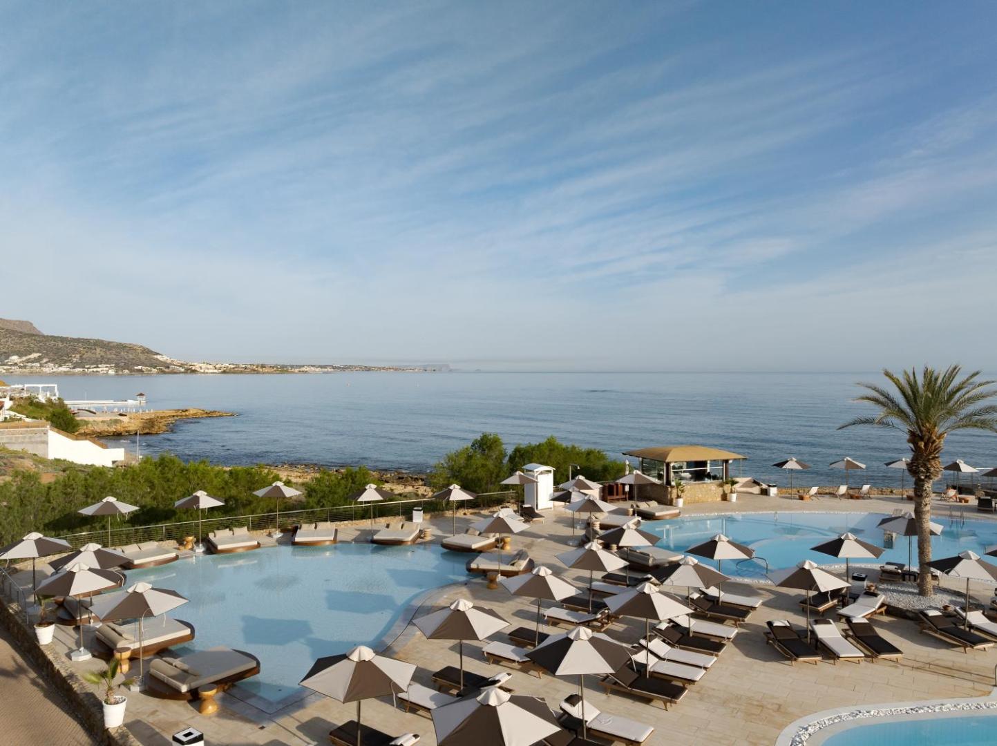Ikaros Beach Resort & Spa galvenais baseins ar skatu uz jūru un kalniem Krētā — plaša sauļošanās zona ar saulessargiem un atpūtas krēsliem