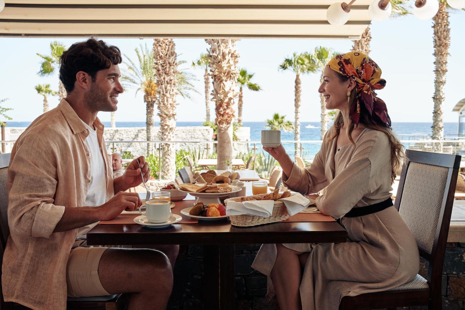 Pāris bauda brokastis Ikaros Beach Resort & Spa āra terasē ar skatu uz palmām un jūru — bagātīgi klāts galds