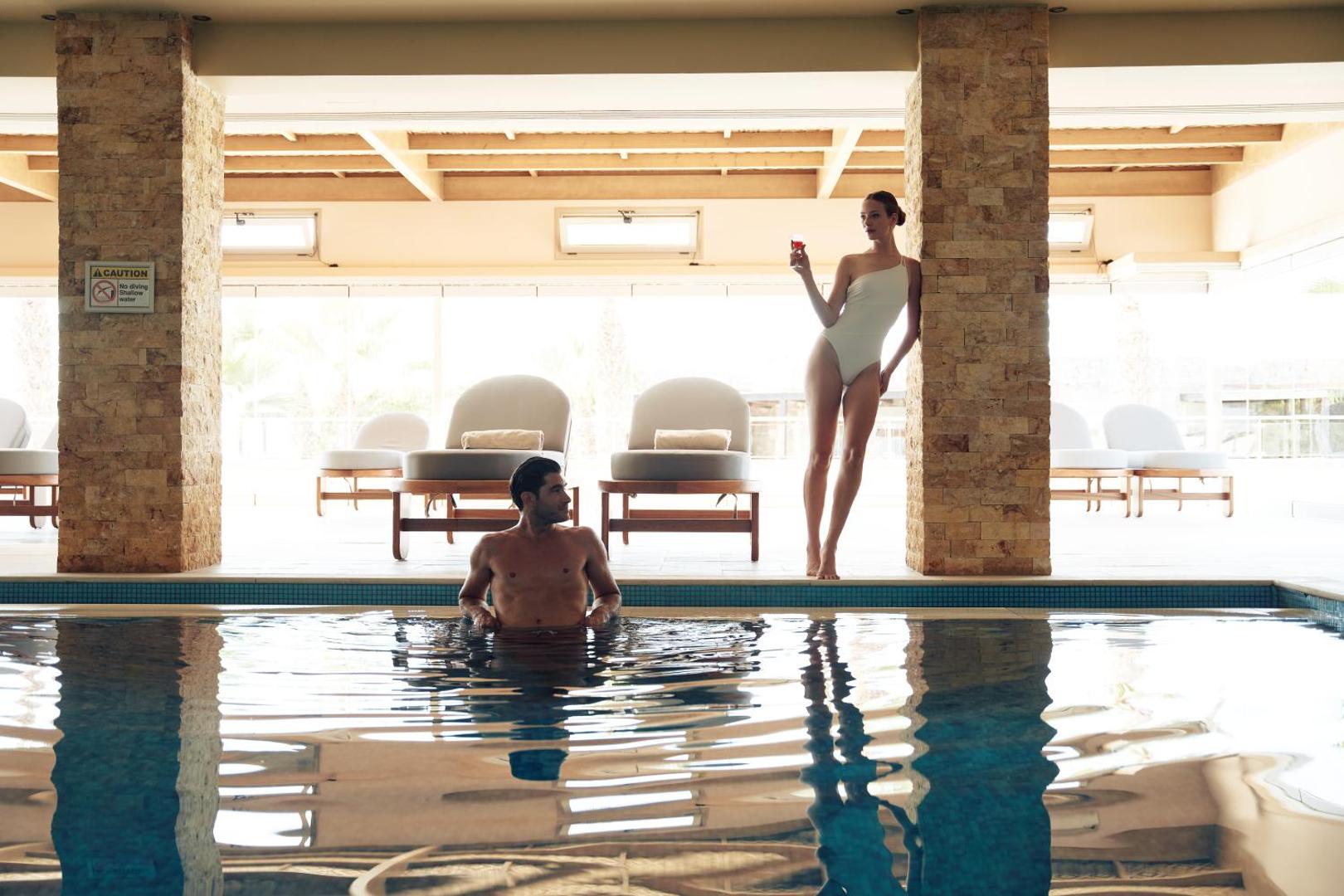 Ikaros Beach Resort & Spa iekštelpu baseins ar atpūtas krēsliem un akmens kolonnām — eleganta atpūtas zona pāriem
