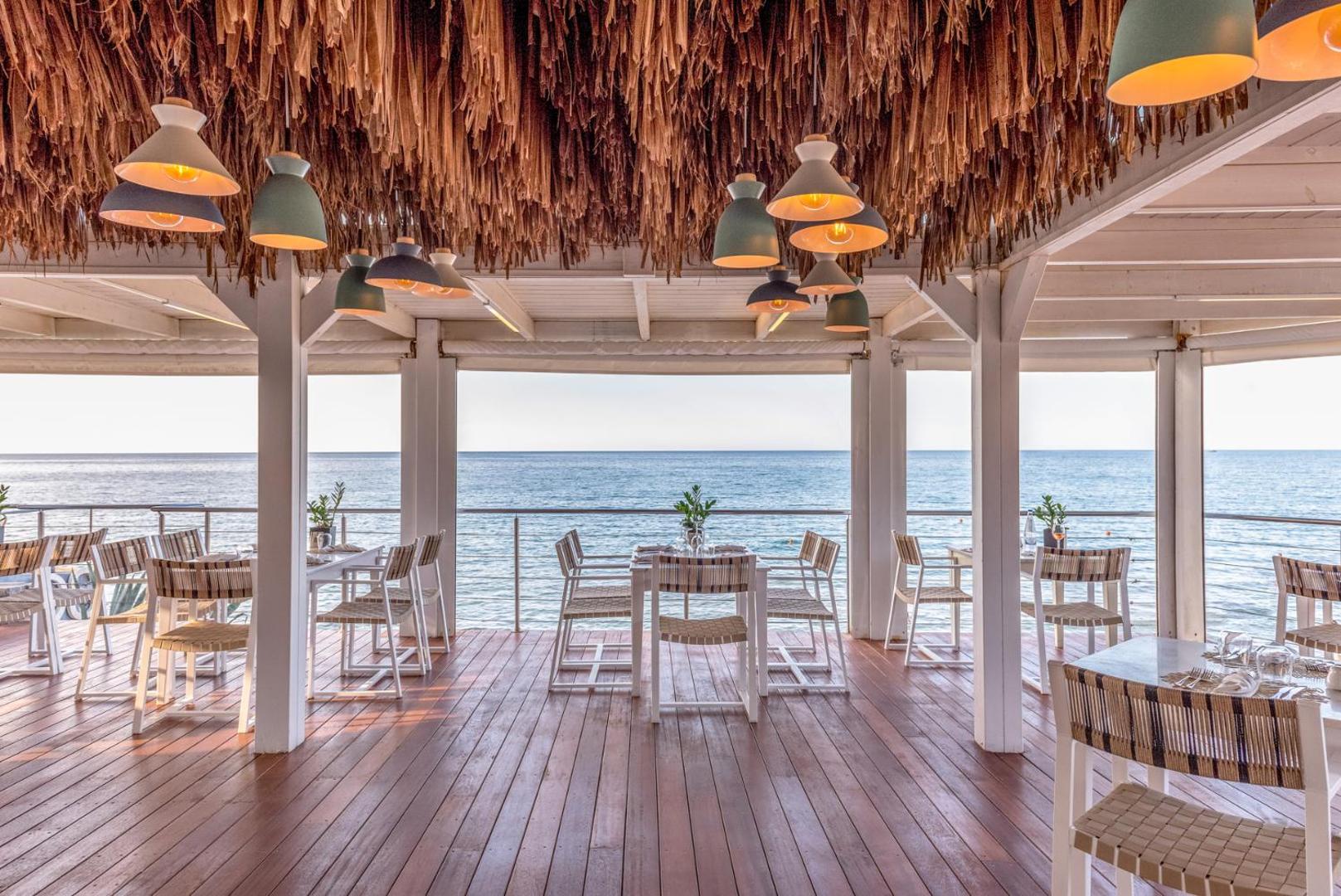 Ikaros Beach Resort & Spa restorāna terase ar koka grīdu un skatu uz jūru — eleganti balti galdi un stilīgs griestu dekors