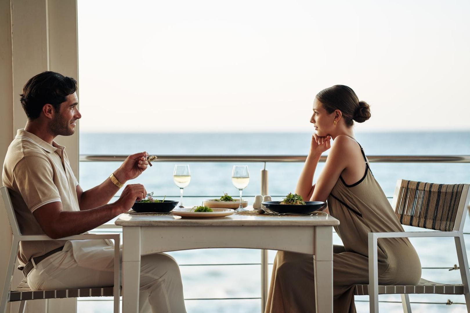 Ikaros Beach Resort & Spa restorāns ar skatu uz jūru — pāris bauda vakariņas uz terases piekrastē saulrieta laikā