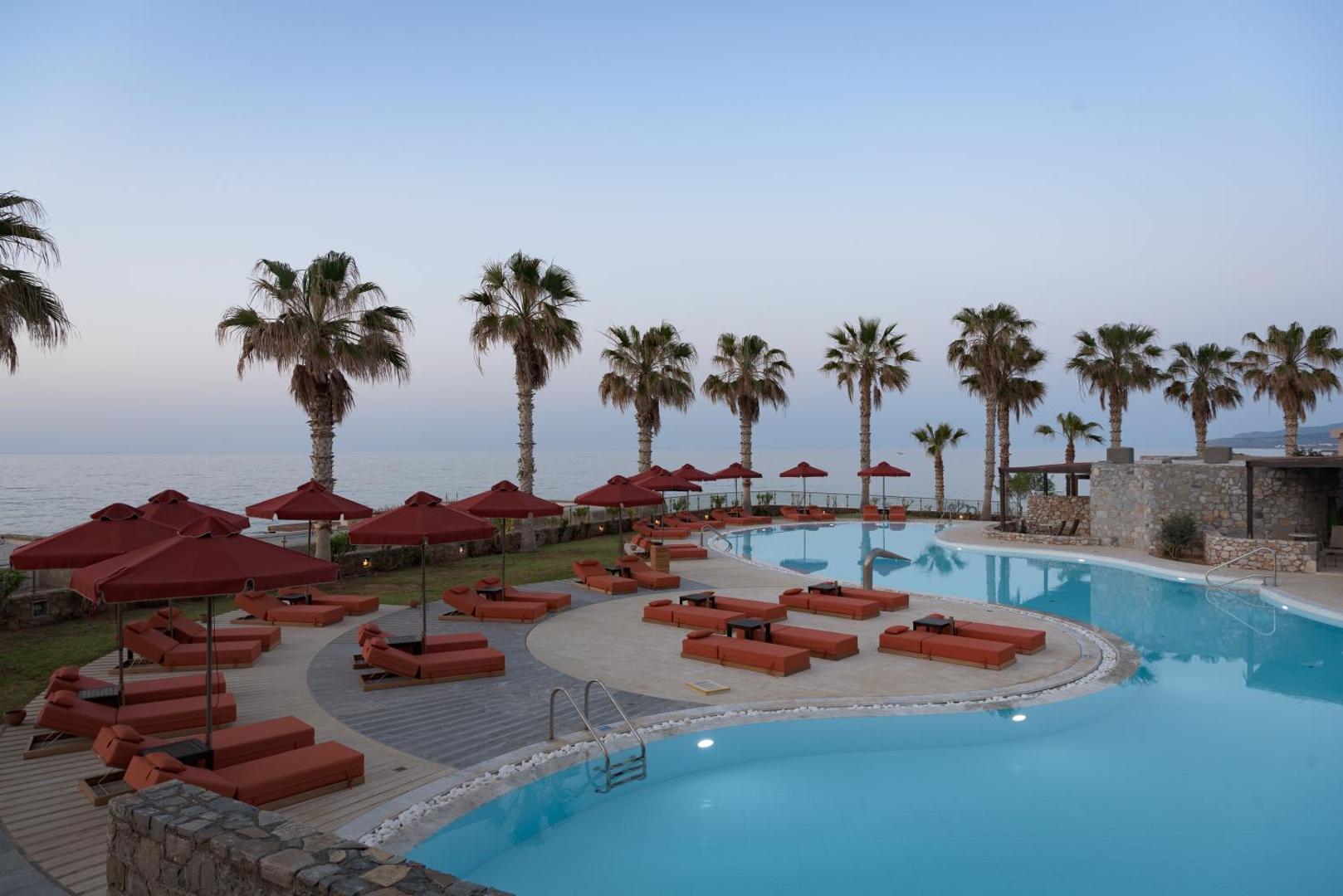 Ikaros Beach Resort & Spa vakara noskaņa pie baseina ar palmām un sarkaniem saulessargiem Krētas piekrastē saulrieta laikā