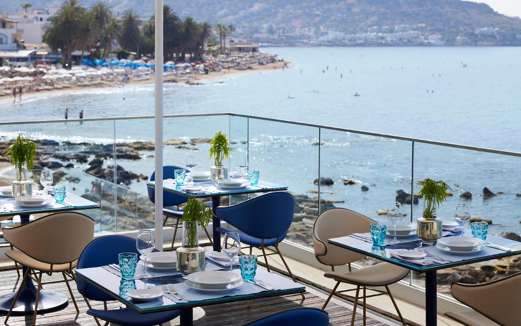 I-Resort Beach Hotel & Spa restorāna terase ar skatu uz jūru un Stalidas piekrasti Krētā — eleganti klāti galdi zilos toņos