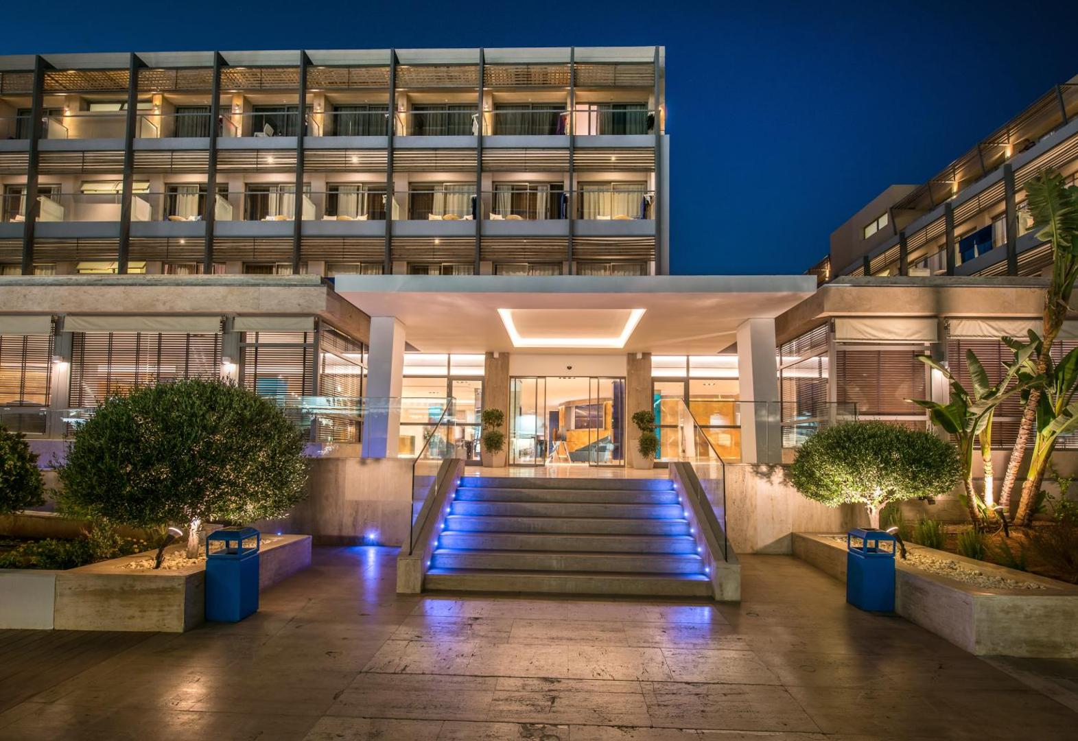 I-Resort Beach Hotel & Spa galvenā ieeja ar izgaismotām kāpnēm un stikla durvīm vakarā — mūsdienīgs dizains un elegants apgaismojums