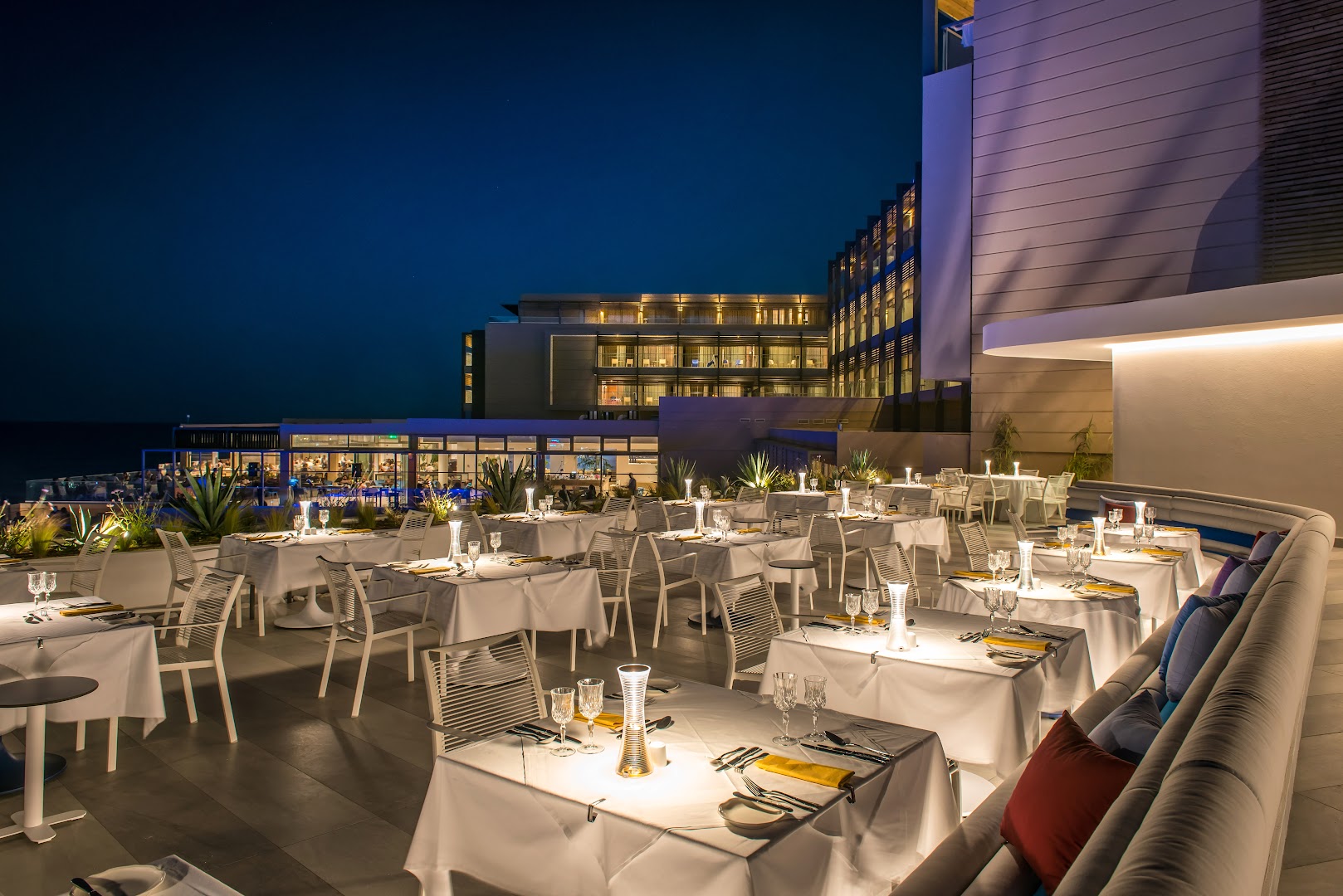 I-Resort Beach Hotel & Spa āra restorāna terase vakarā ar modernu apgaismojumu un skatu uz viesnīcas galveno ēku