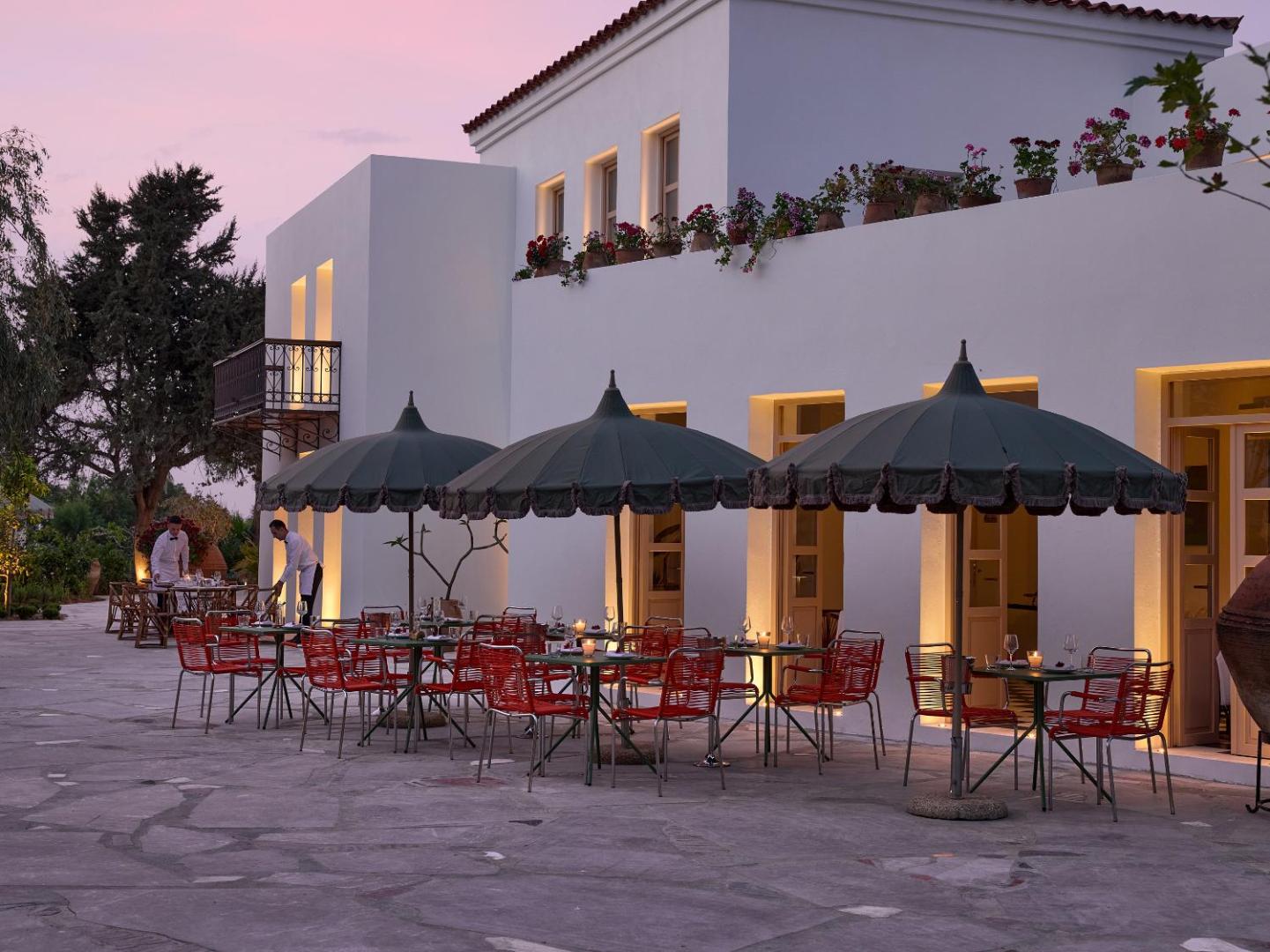 Grecotel Creta Palace Resort restorāna āra terase krēslas stundā ar sarkaniem krēsliem, saulessargiem un izgaismotu viesnīcas fasādi Krētā