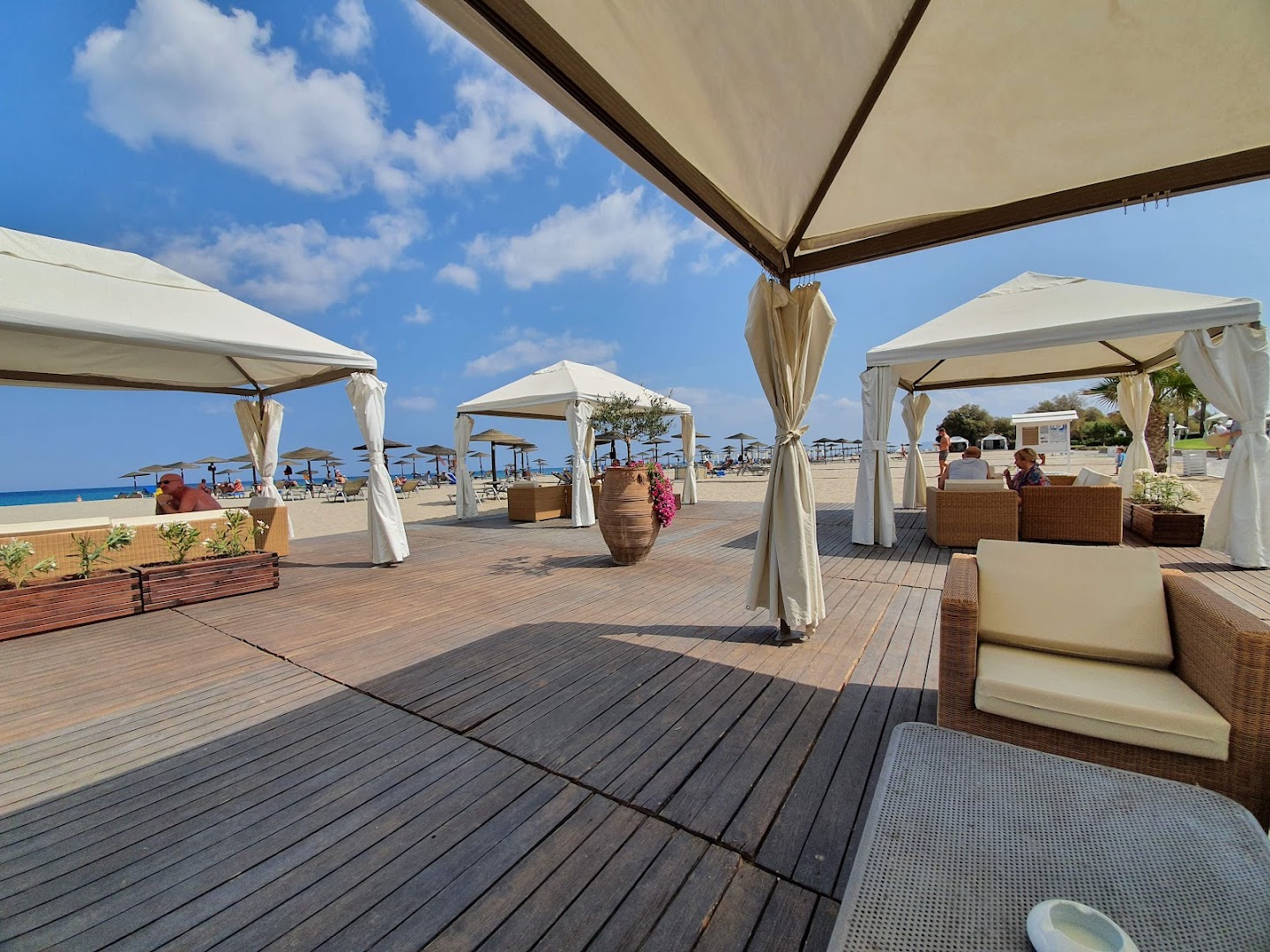 Grecotel Creta Palace Resort pludmales atpūtas zona ar koka klāju, baltām teltīm un ērtiem dīvāniem pie smilšu krasta