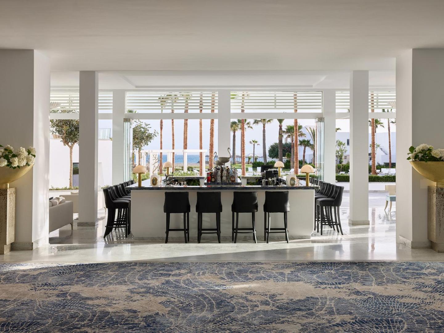 Grecotel Creta Palace Resort lobby bārs ar skatu uz palmu dārzu un jūru, gaišs interjers ar augstiem griestiem