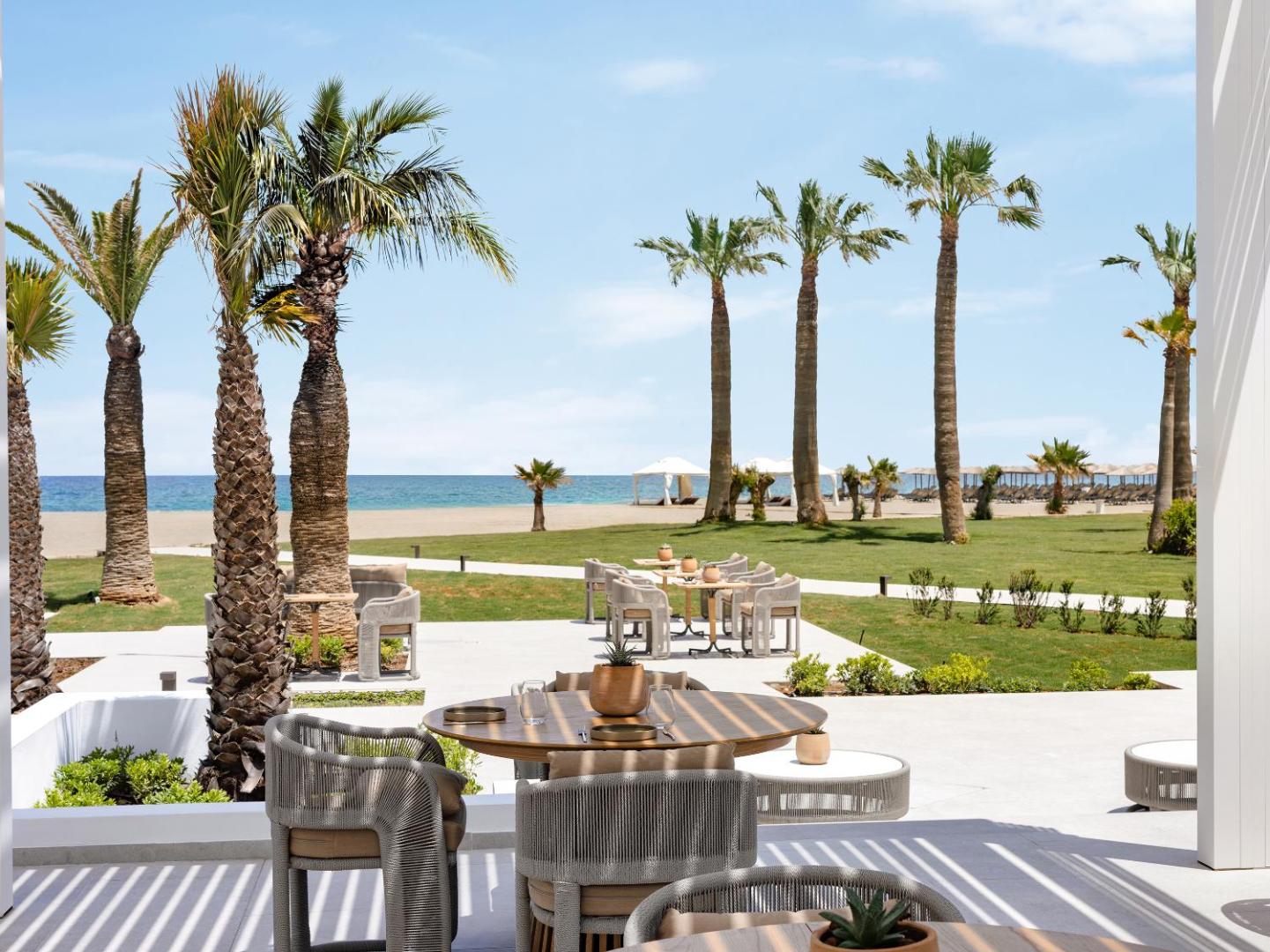 Grecotel Creta Palace Resort restorāna zona ar skatu uz jūru, palmām un smilšu pludmali saulainā dienā Krētas piekrastē