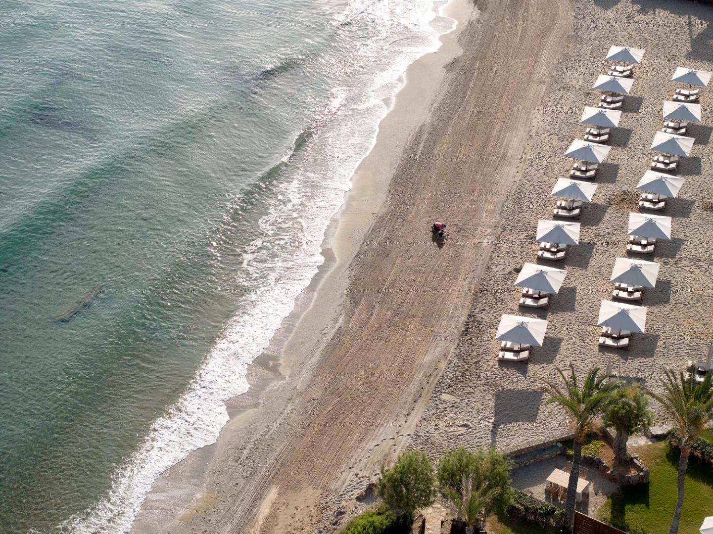 Grecotel Amirandes Exclusive Resort plašā smilšu pludmale ar saulessargiem un zviļņiem — skats no putna lidojuma uz dzidro jūru