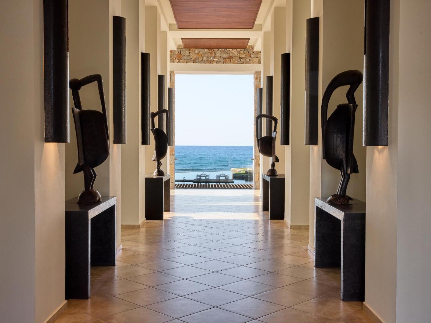 Grecotel Amirandes viesnīcas gaitenis ar modernām skulptūrām un skatu uz zilo Vidusjūru un baseinu Krētas piekrastē