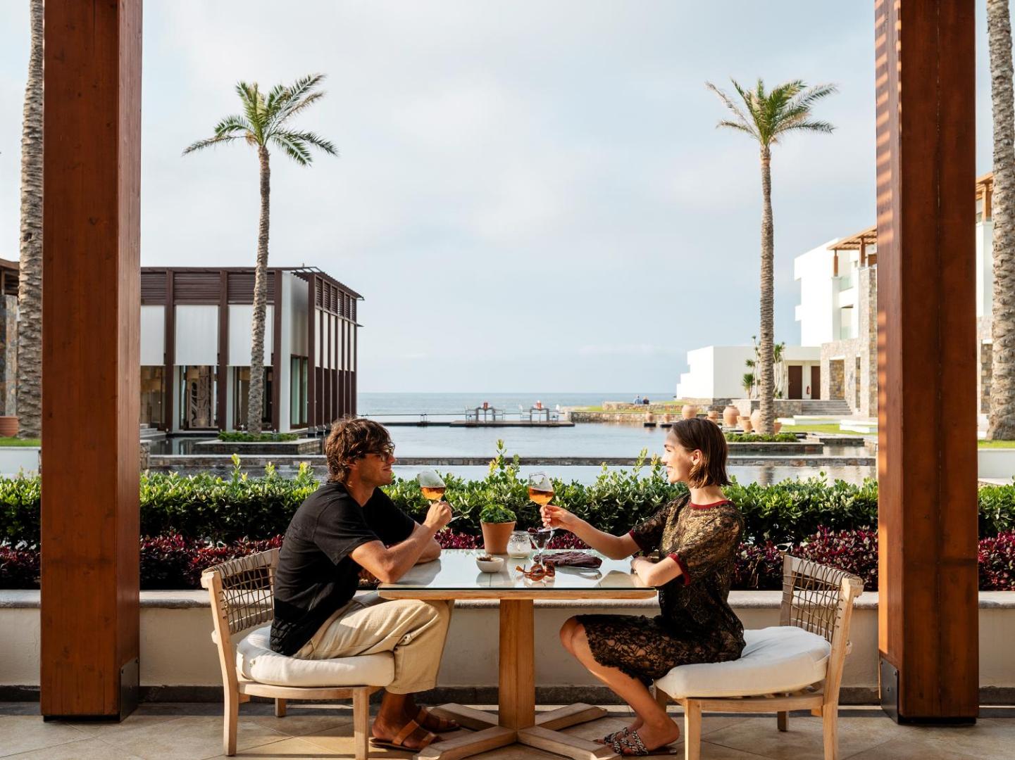 Pāris bauda dzērienus Grecotel Amirandes restorāna āra terasē — fonā palmas un viesnīcas modernā arhitektūra pie ūdens