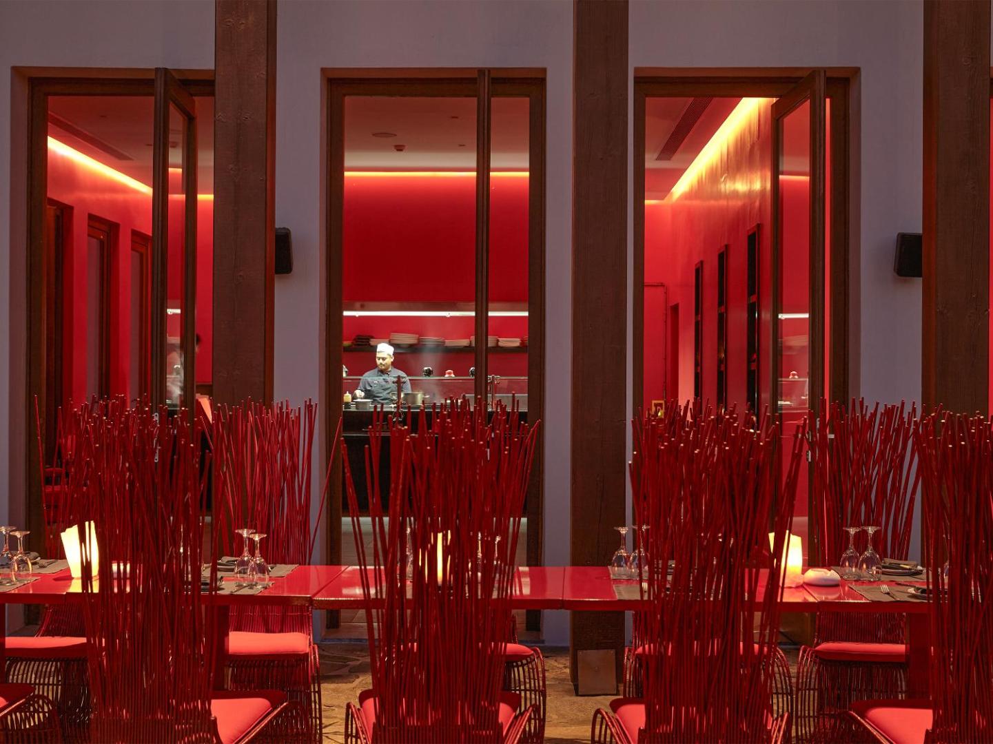 Grecotel Amirandes restorāns Minotaur ar koši sarkanu interjeru, dizaina krēsliem un atvērto virtuvi, kur redzams šefpavārs darbībā