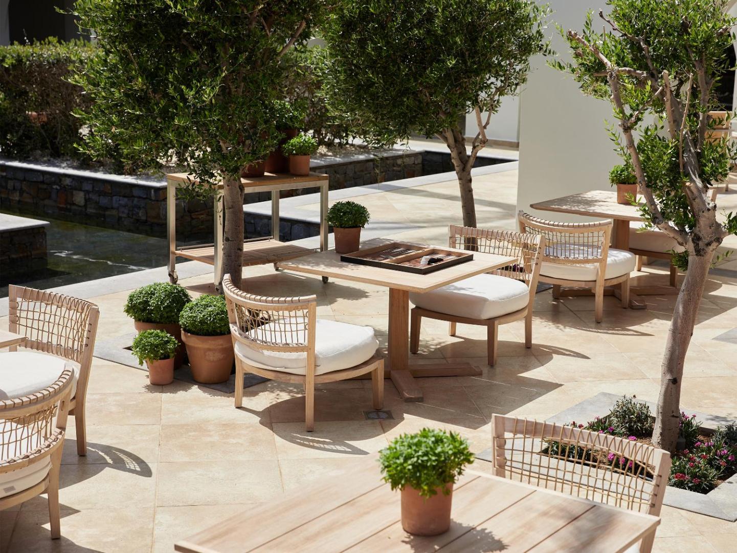 Grecotel Amirandes Exclusive Resort āra terase ar koka galdiem un pītiem krēsliem olīvkoku pavēnī, kur pieejamas galda spēles viesu atpūtai