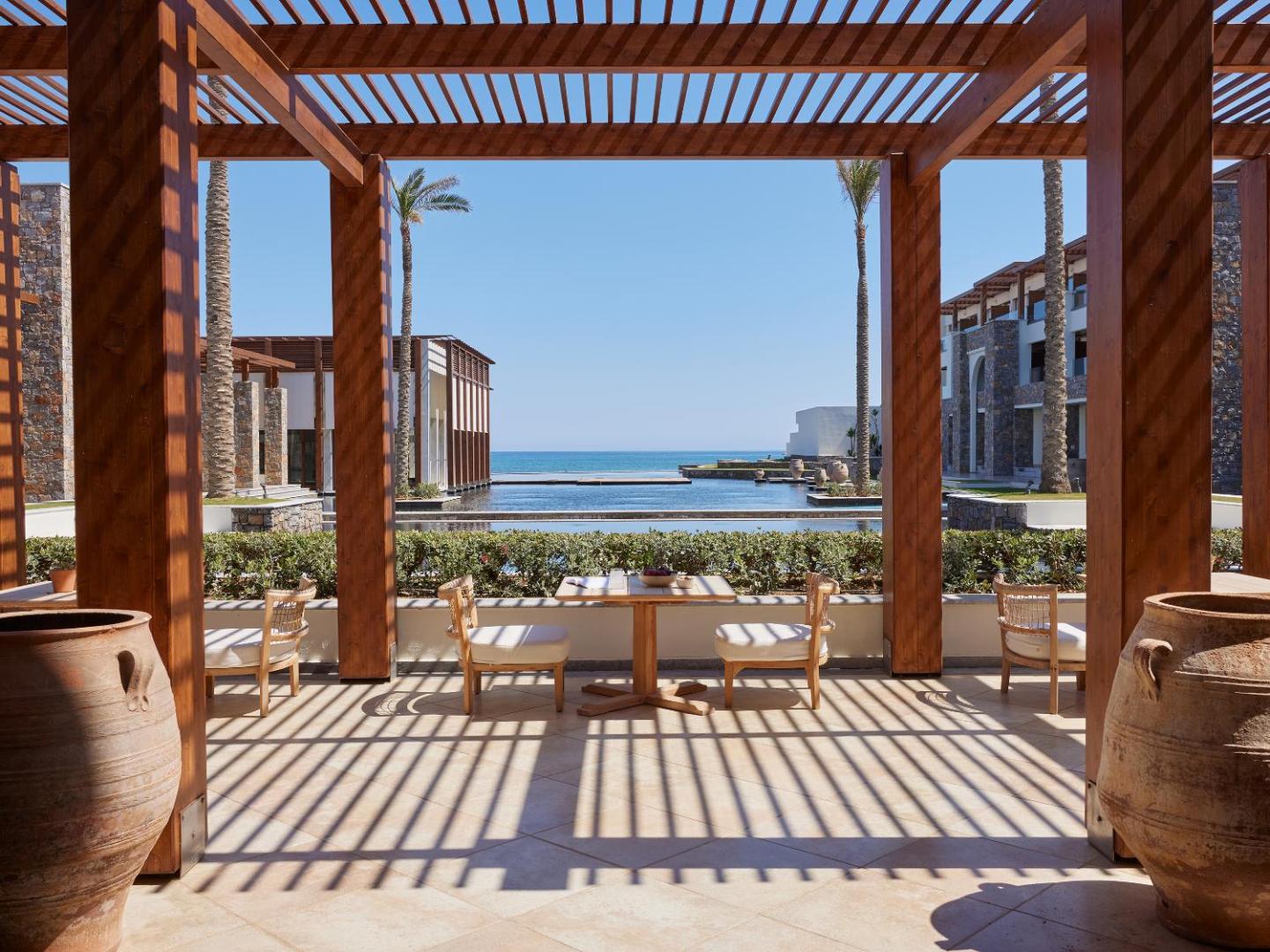Grecotel Amirandes restorāna terase ar koka pergolu un skatu uz dekoratīvajiem baseiniem un jūru — elegants galdiņš diviem