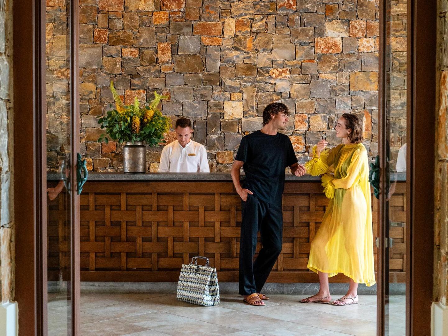 Grecotel Amirandes reģistratūras zona ar akmens sienu un koka apdari, kur viesmīlīgs personāls uzņem viesus pie ierašanās