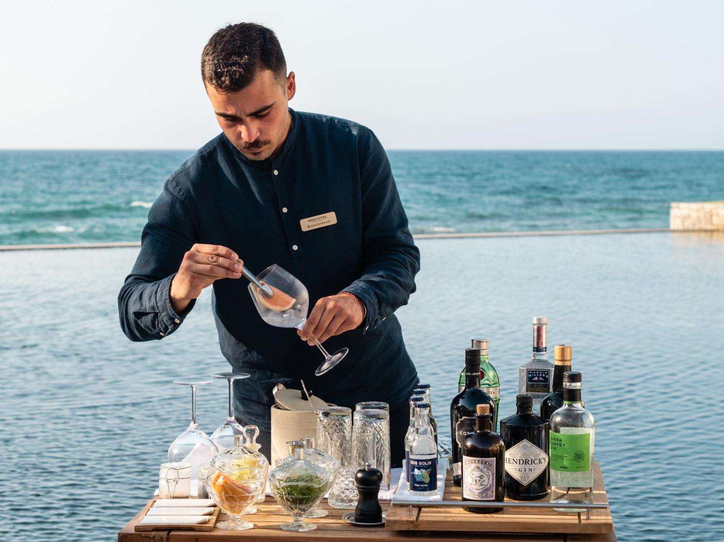 Grecotel Amirandes bārmenis gatavo kokteili pie āra baseina Krētā — fona redzama jūra un plašs džina klāsts uz koka galda