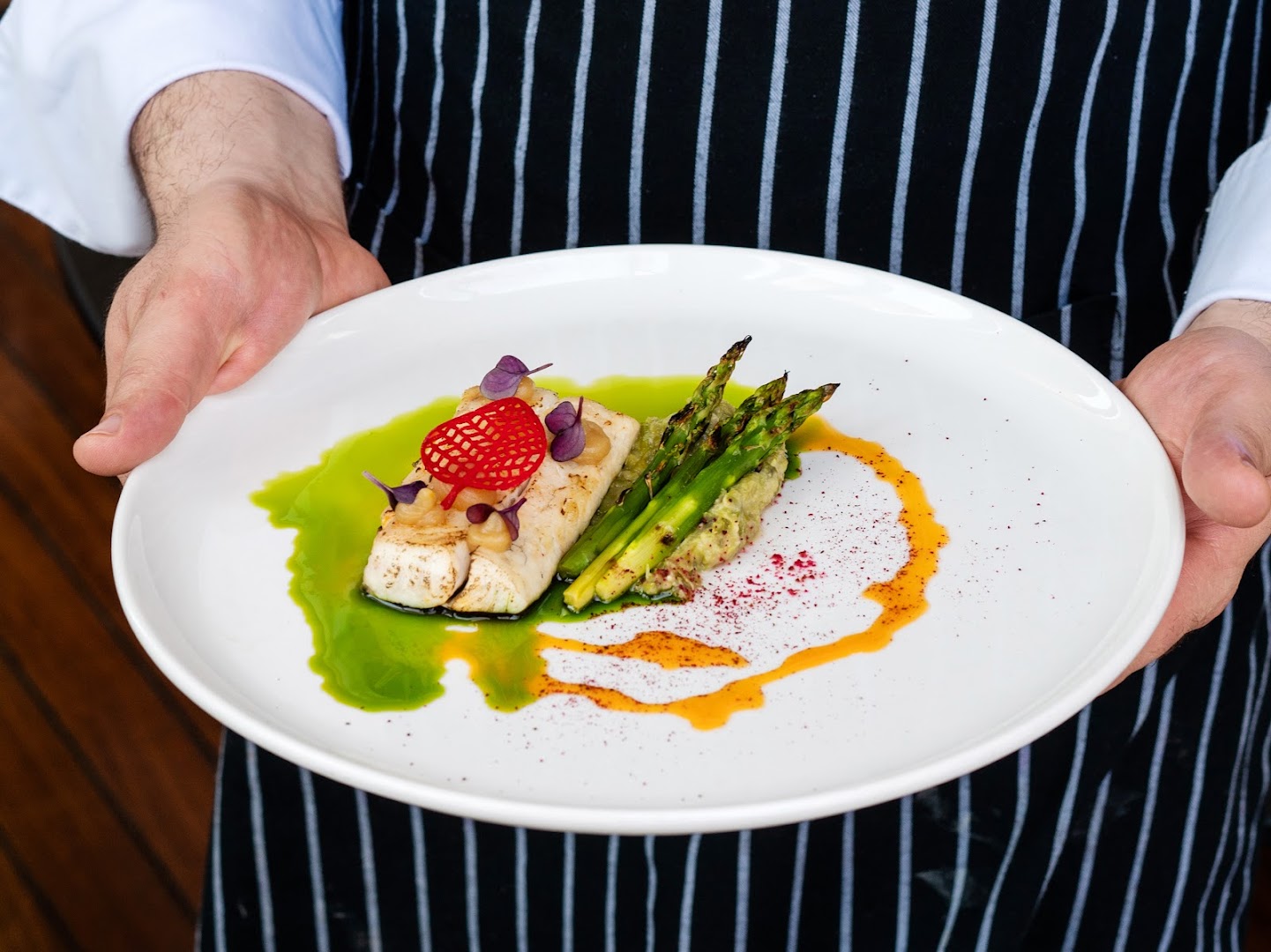 Grecotel Amirandes šefpavārs pasniedz grilētu zivi ar sparģeļiem un biezeni uz balta šķīvja — izsmalcināta restorāna virtuve Krētā