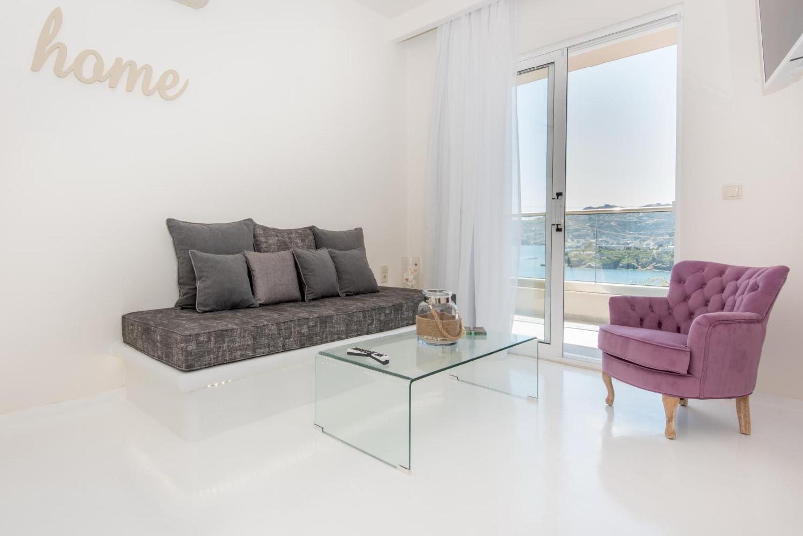 Eva Mare Hotel & Suites viesistabas zona ar pelēku dīvānu, violetu atpūtas krēslu un stikla galdiņu pie plaša loga