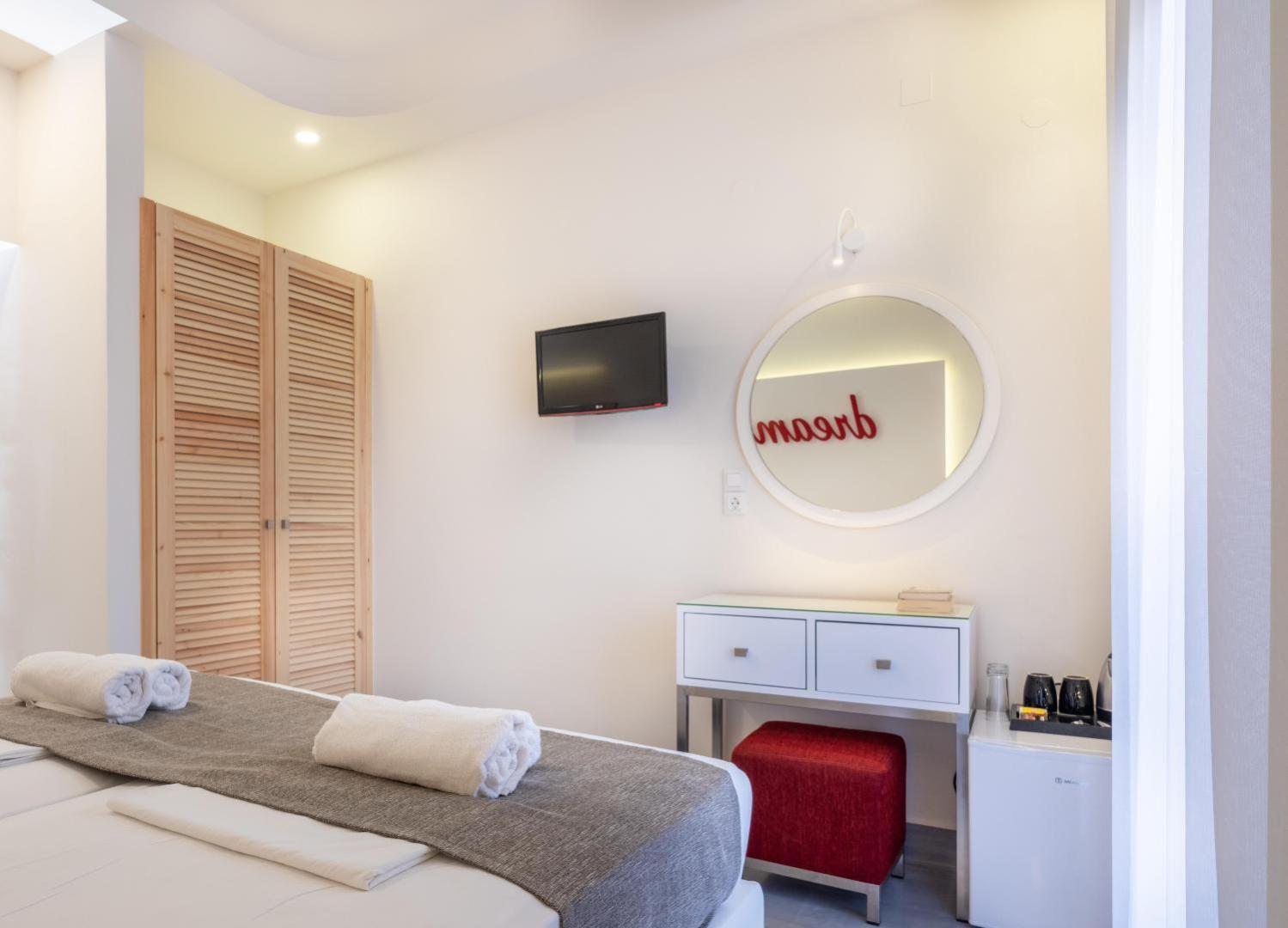 Eva Mare Hotel & Suites numura interjers ar baltu tualetes galdiņu, apaļu spoguli, sarkanu pufu un koka drēbju skapi