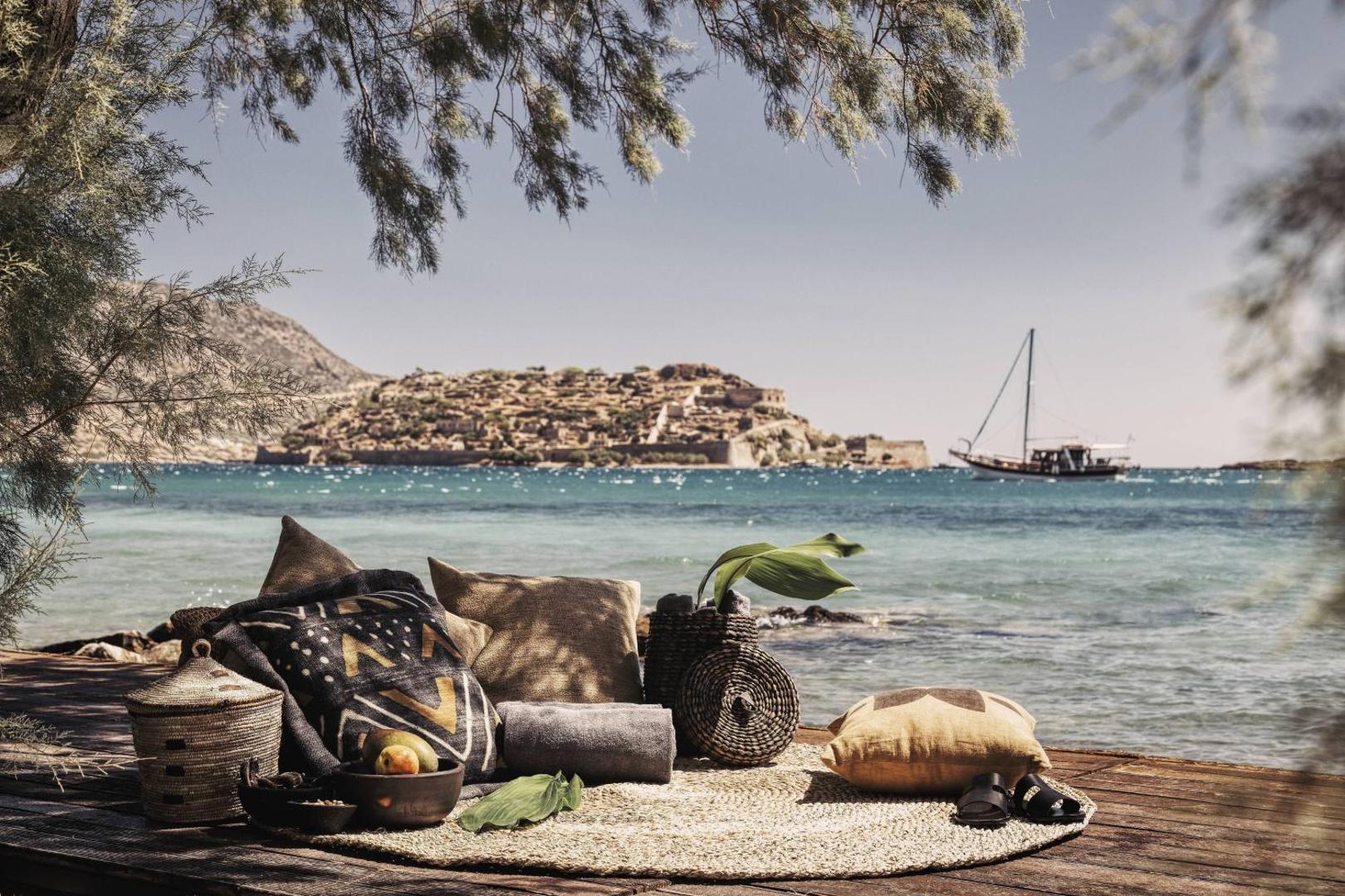 Domes of Elounda pludmales atpūtas zona ar paklāju, spilveniem un groziem koku pavēnī, fonā jūra un buru laiva