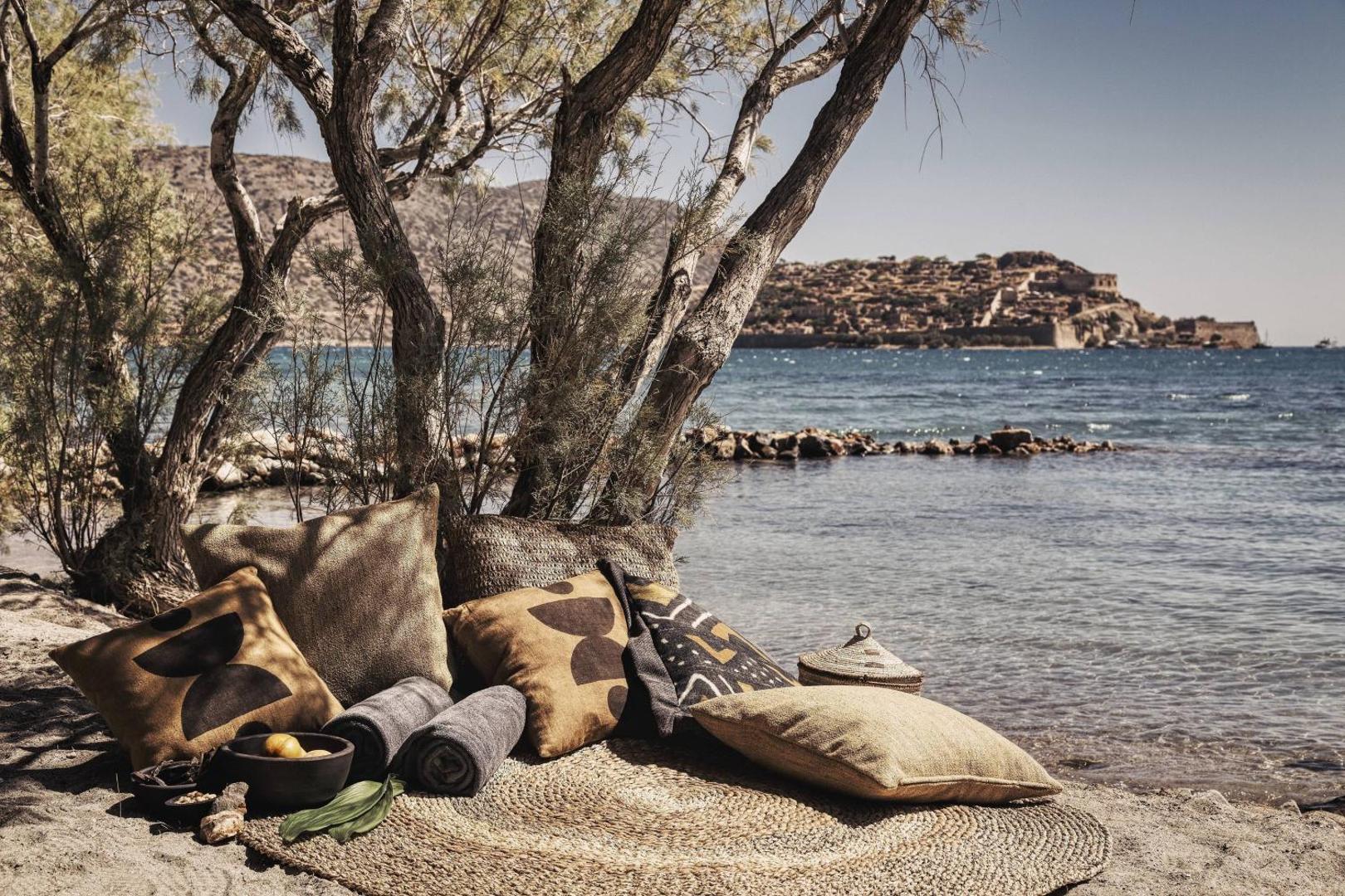 Mājīga atpūtas vieta Domes of Elounda pludmales smiltīs ar mīkstiem spilveniem un dvieļiem koku ēnā pie jūras