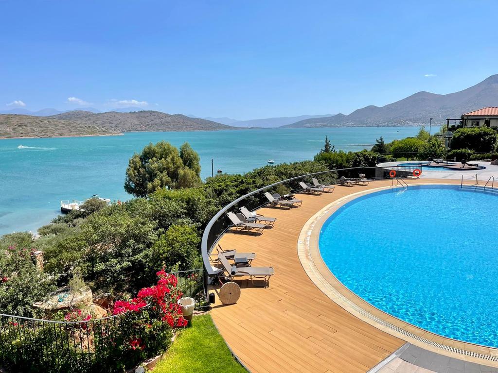 Domes of Elounda Autograph Collection apaļas formas baseins ar koka klāju un sauļošanās krēsliem piekrastē ar skatu uz kalniem