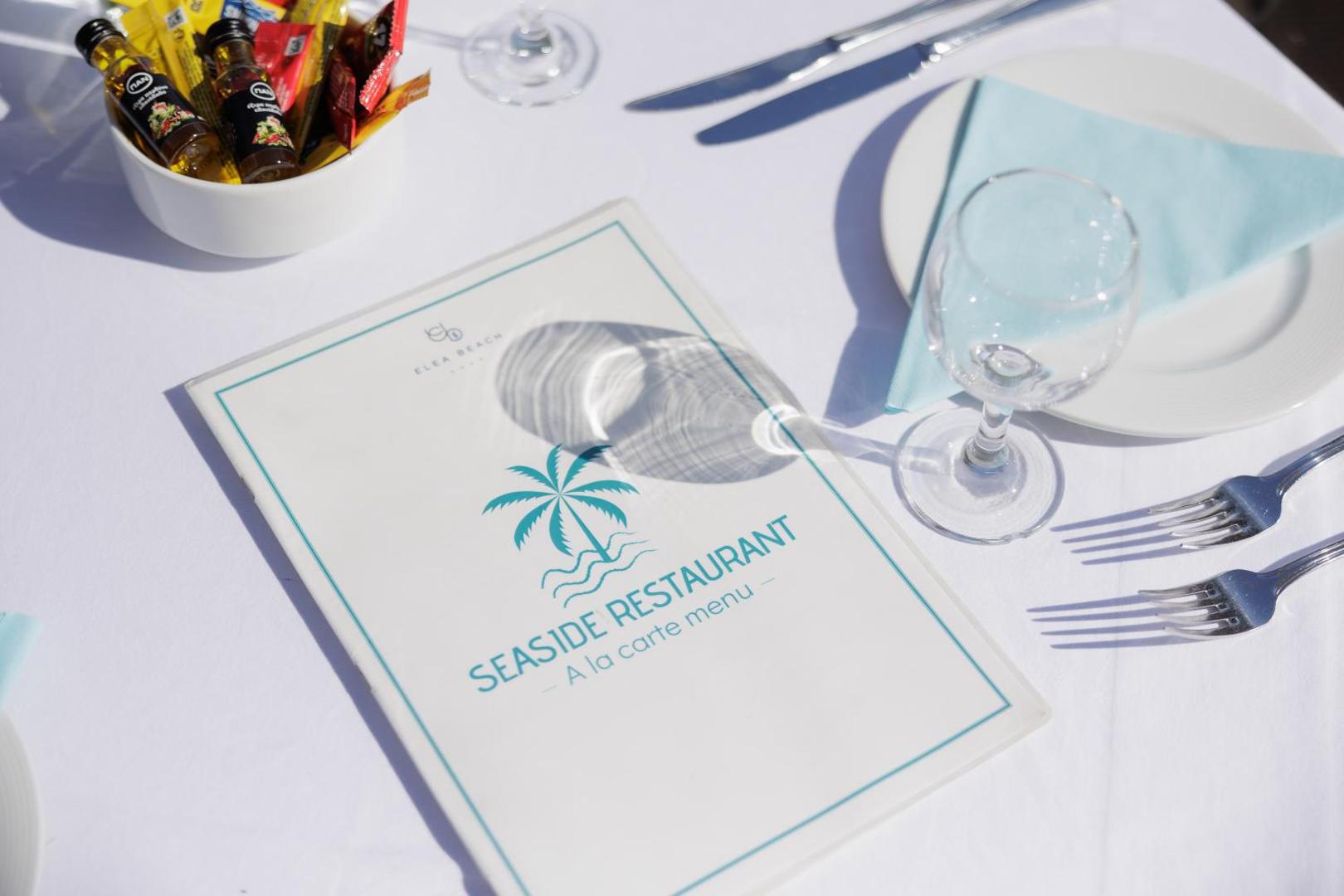 Elea Beach Hotel Seaside ēdienkartes restorāna a la carte menu uz balta galdauta ar vīna glāzi un galda piederumiem