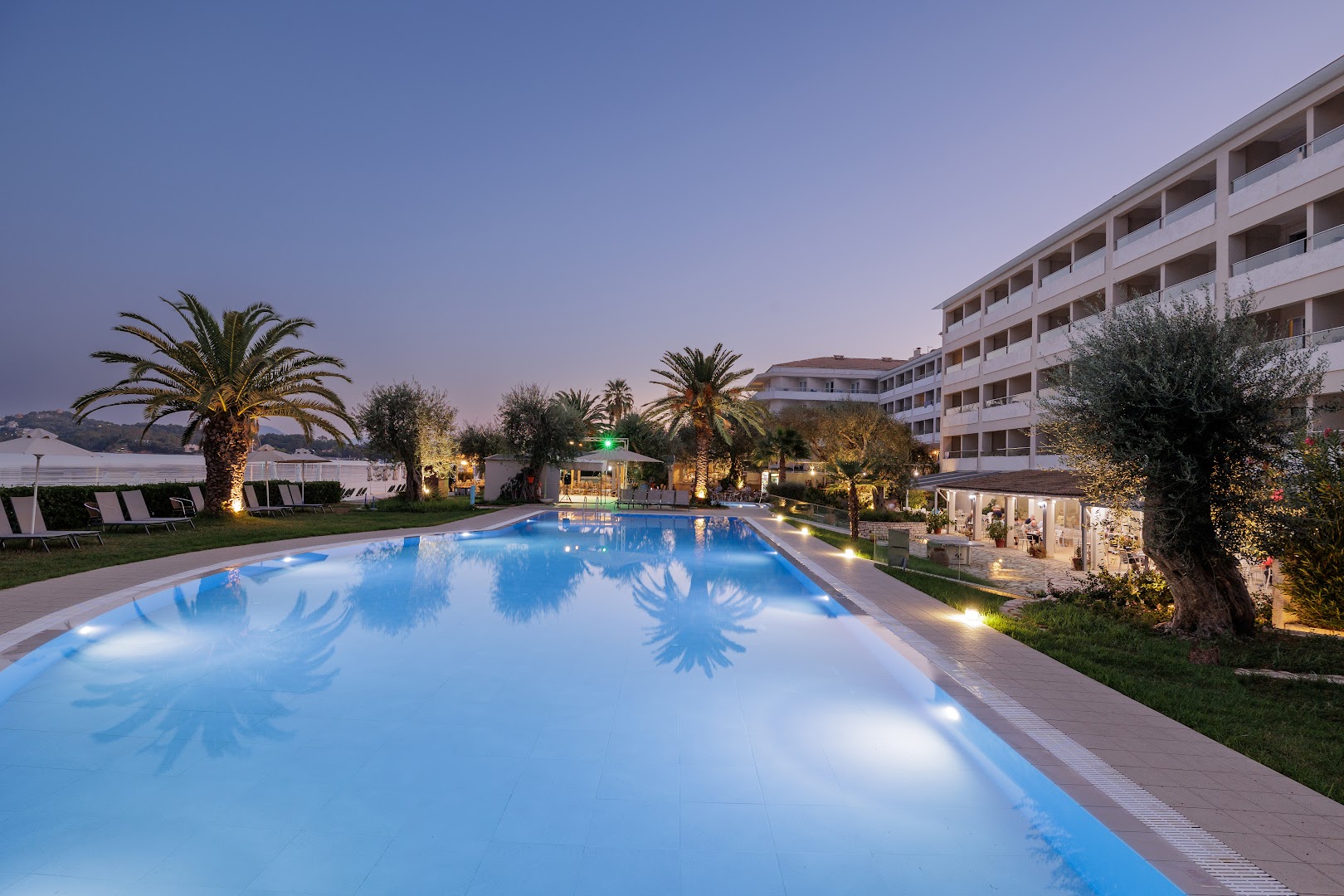 Elea Beach Hotel izgaismots āra baseins vakarā pie viesnīcas korpusa Dasijā — vakara atmosfēra ar palmām un terasi