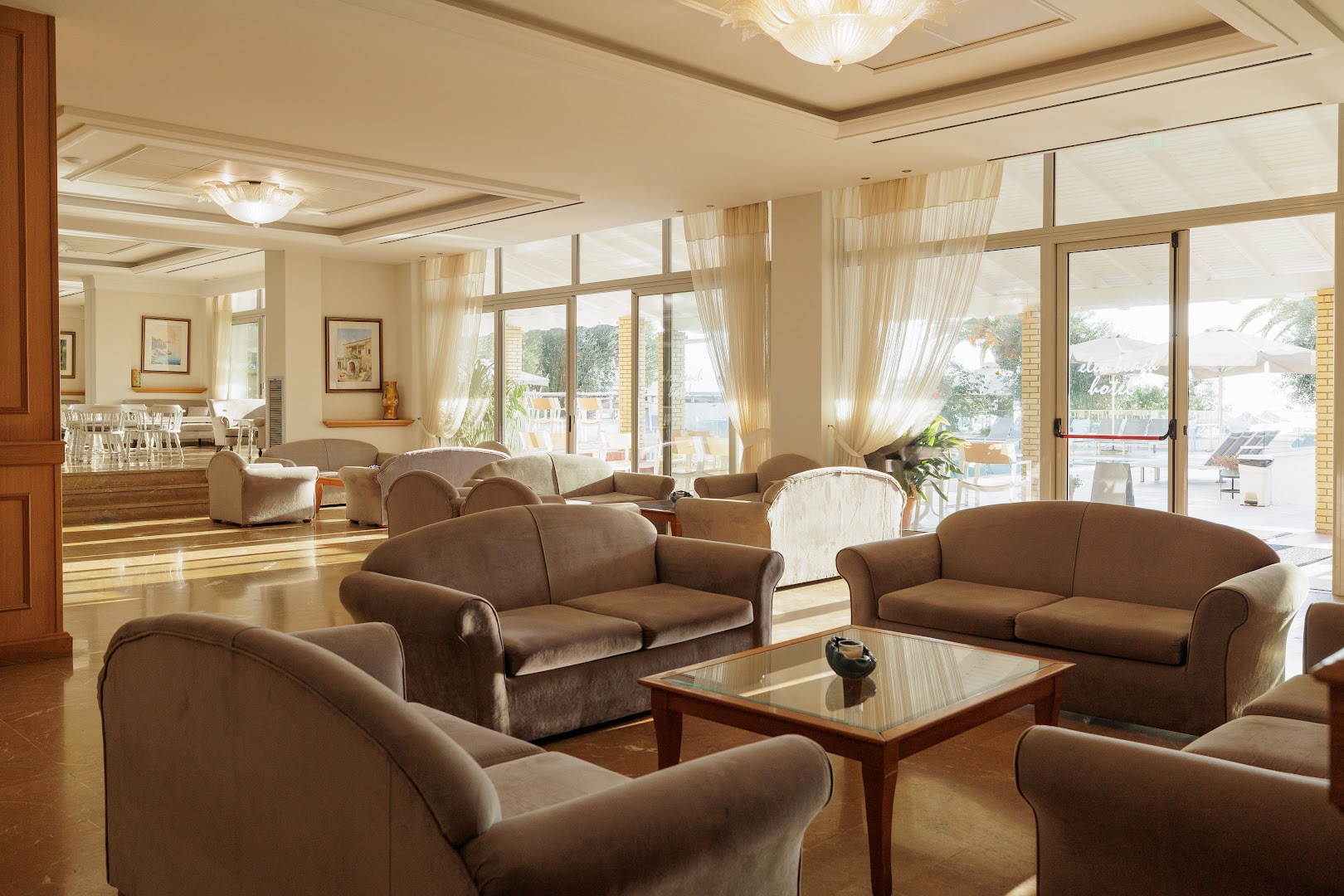 Elea Beach Hotel plašā lobby atpūtas zona ar mīkstajiem dīvāniem, stikla galdiņiem un lieliem logiem, kas vērsti uz terasi