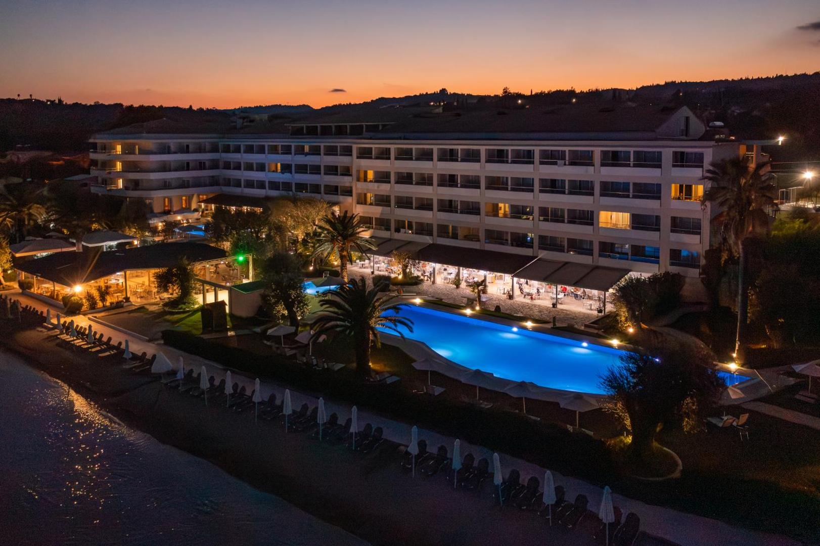 Elea Beach Hotel izgaismotais baseins un viesnīcas fasāde vakarā ar āra terasi un saulrieta debesīm